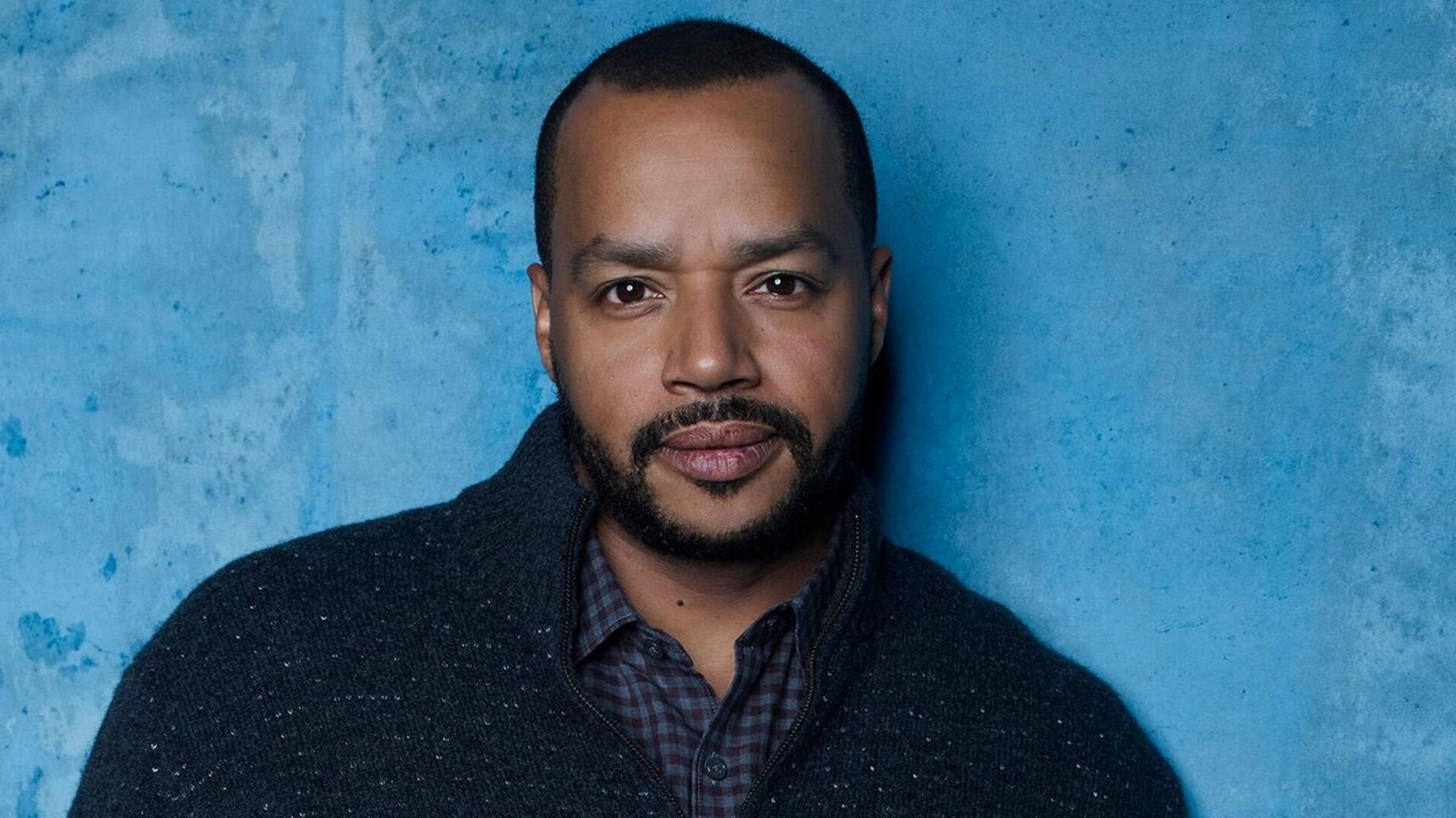 Aktor Donald Faison w niebieskim swetrze na jasno niebieskim tle