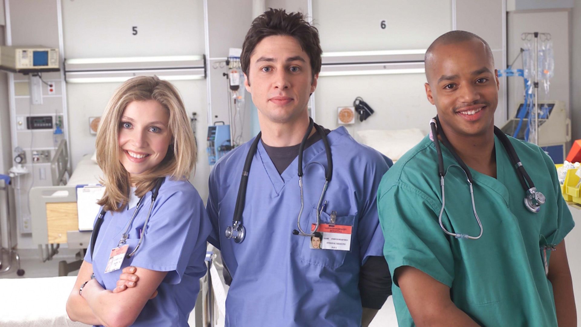 Sarah Chalke, Zach Braff i Donald Faison w medycznych fartuchach stoją w szpitalnym pokoju i się uśmiechają