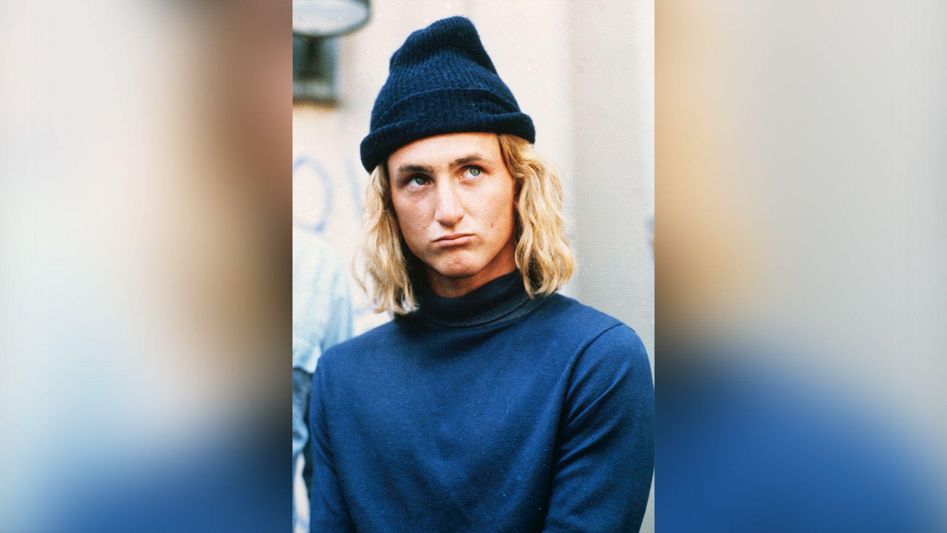 Młody Sean Penn z długimi włosami, ubrany w niebieski golf i niebieską czapkę