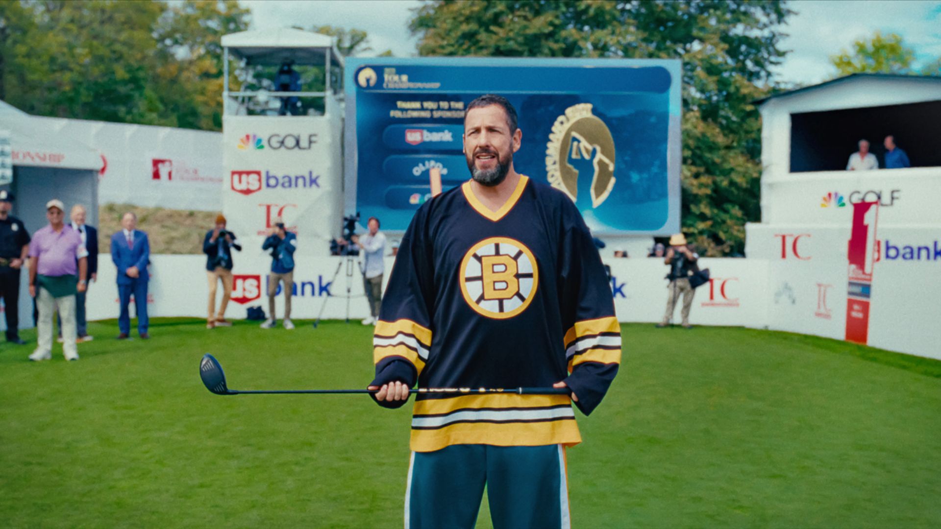 Adam Sandler w koszulce trzyma kij hokejowy, stojąc na stadionie