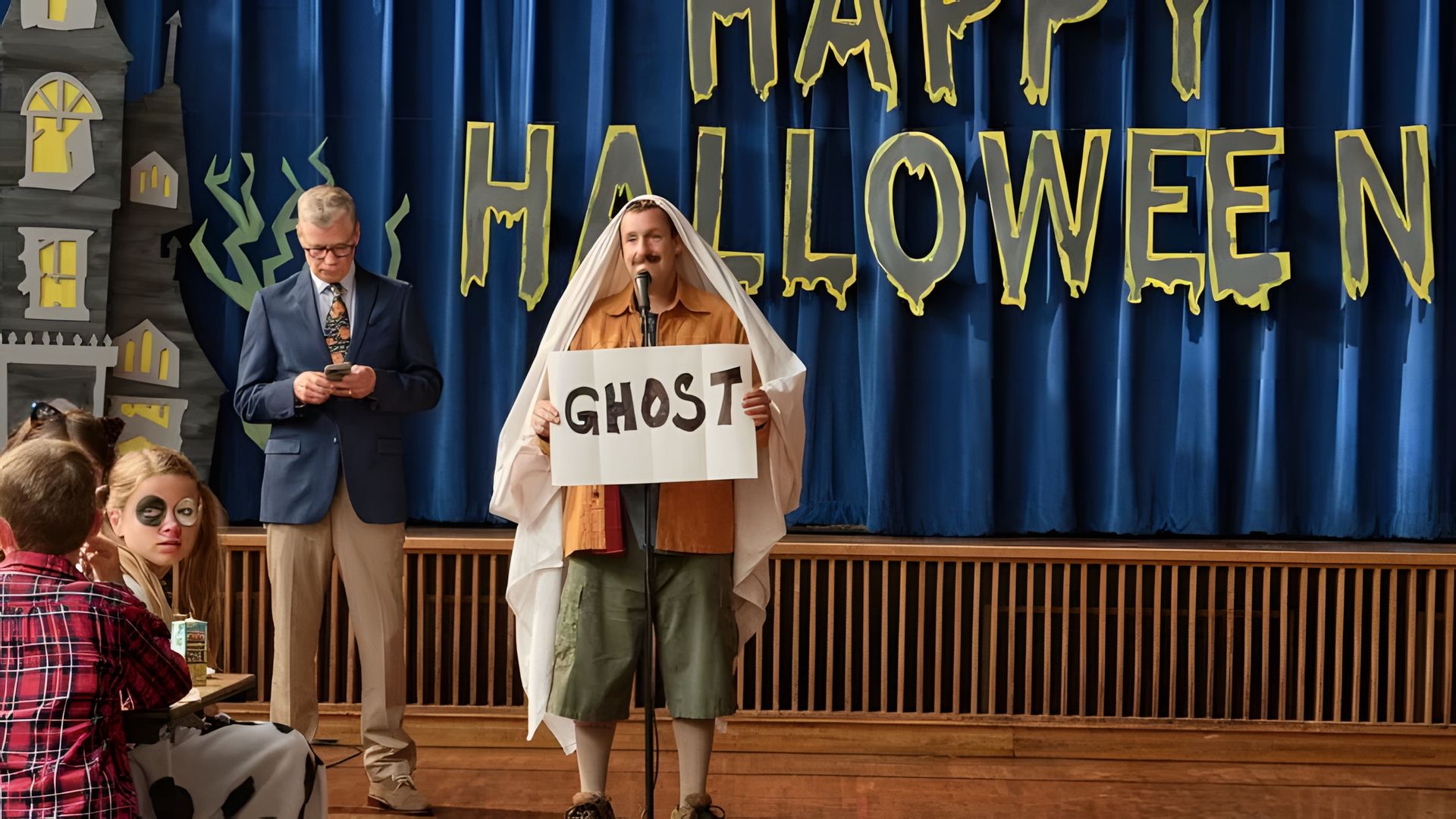 Adam Sandler w prześcieradle na głowie, trzymający tabliczkę z napisem GHOST