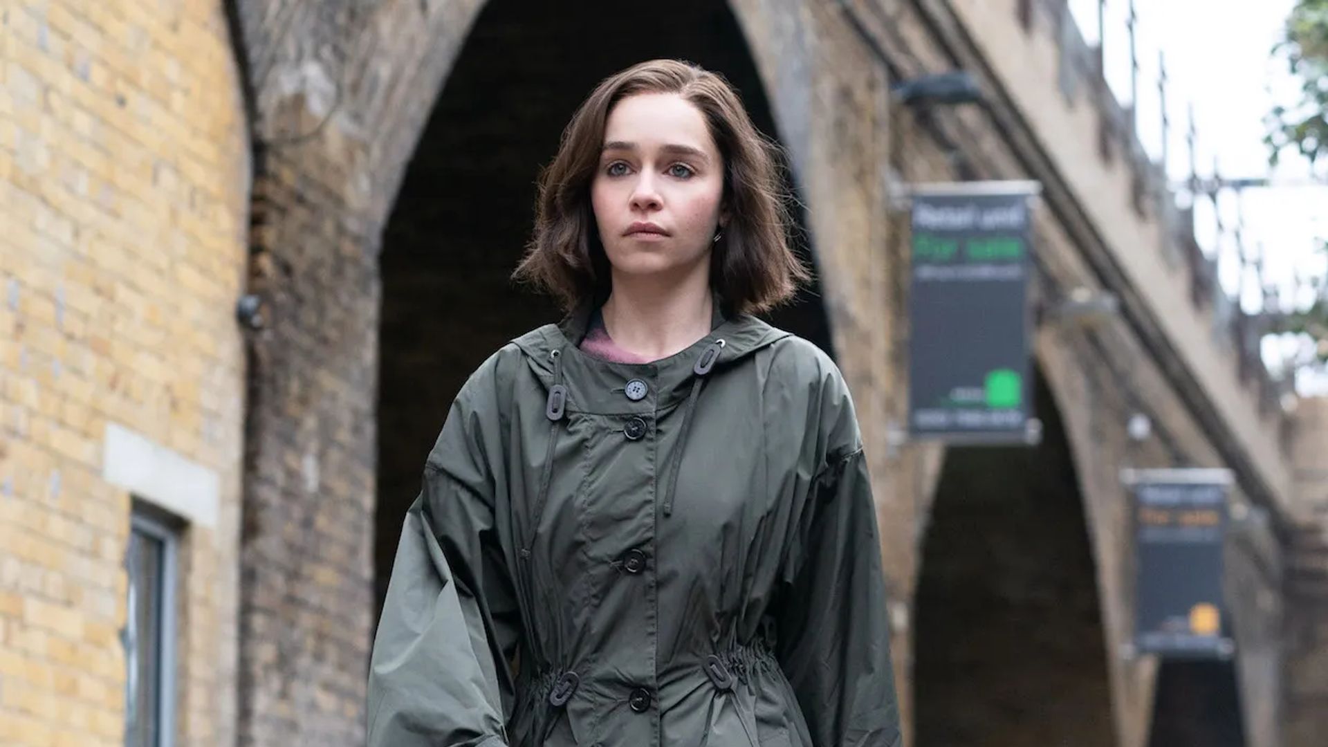 Emilia Clarke w płaszczu przeciwdeszczowym idzie ulicą, skupione spojrzenie przed siebie