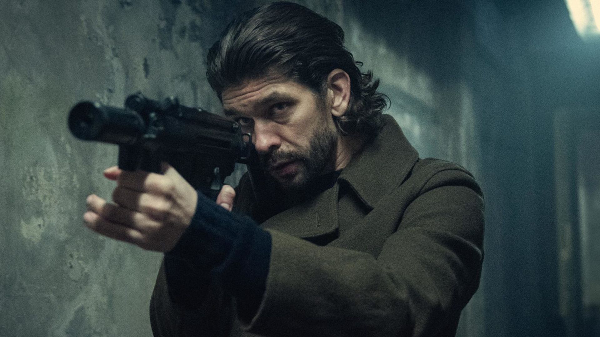 Ben Whishaw z długimi włosami w płaszczu trzymający broń