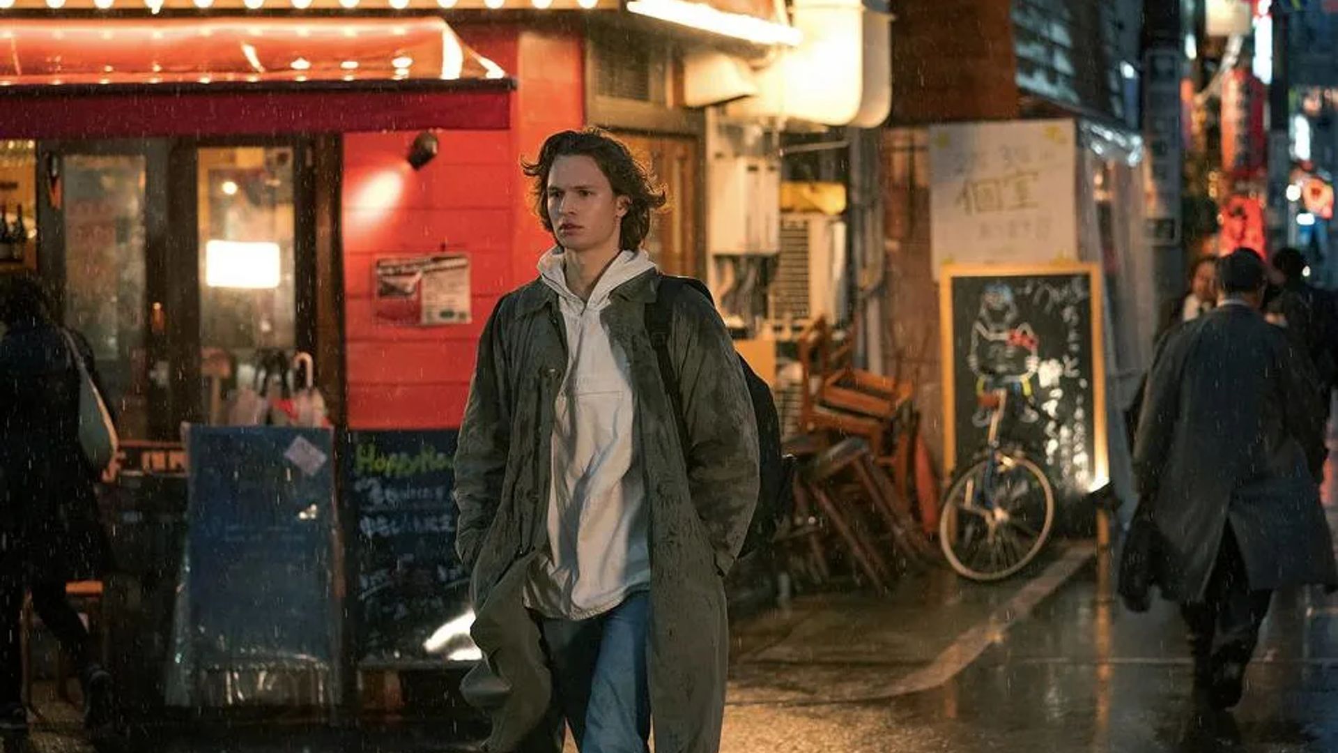 Ansel Elgort w płaszczu i bluzie z kapturem idzie deszczową ulicą obok jasno oświetlonych witryn sklepowych i restauracji