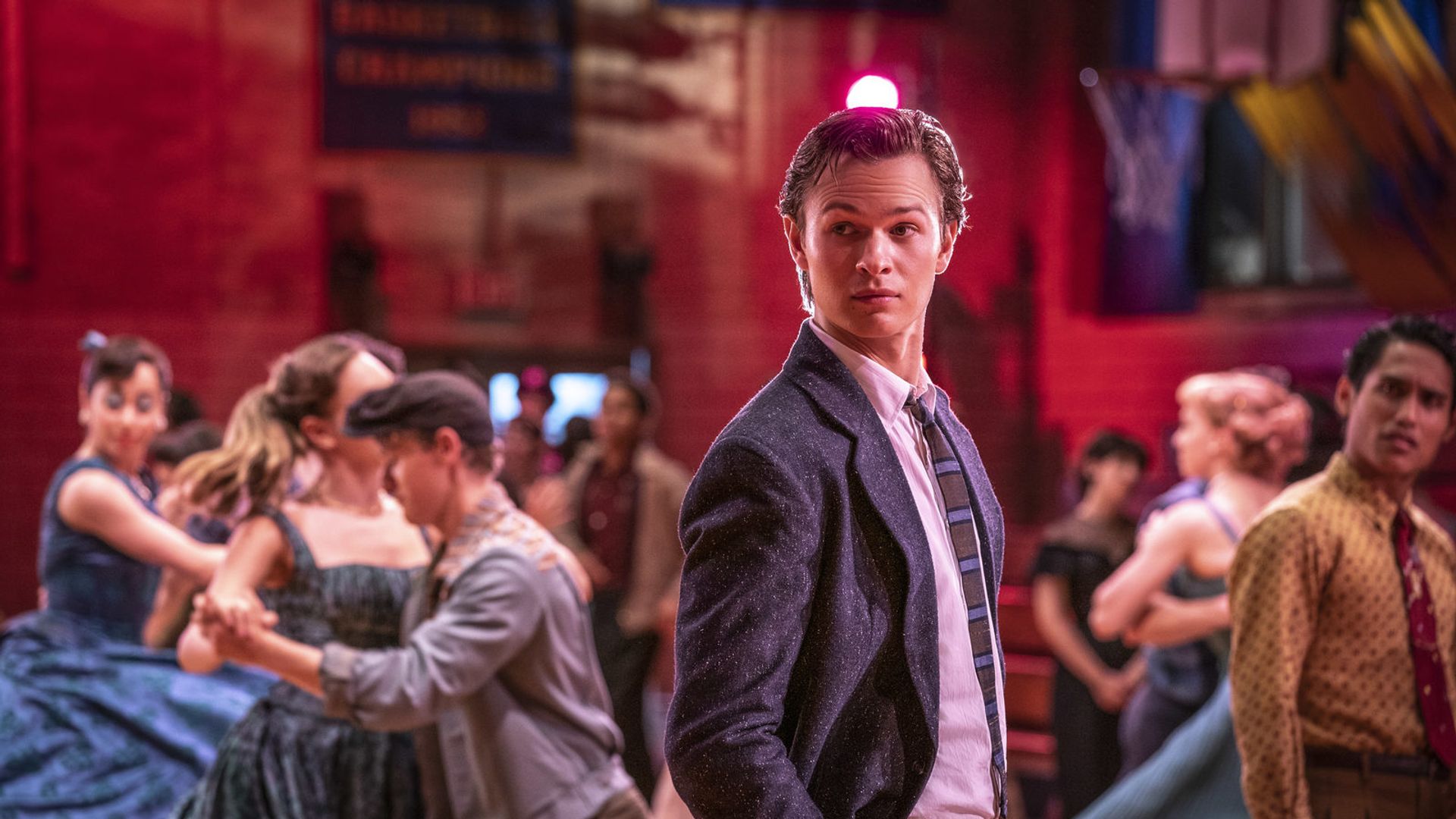 Ansel Elgort w garniturze i krawacie stoi na szkolnej potańcówce, wokół niego wirują pary