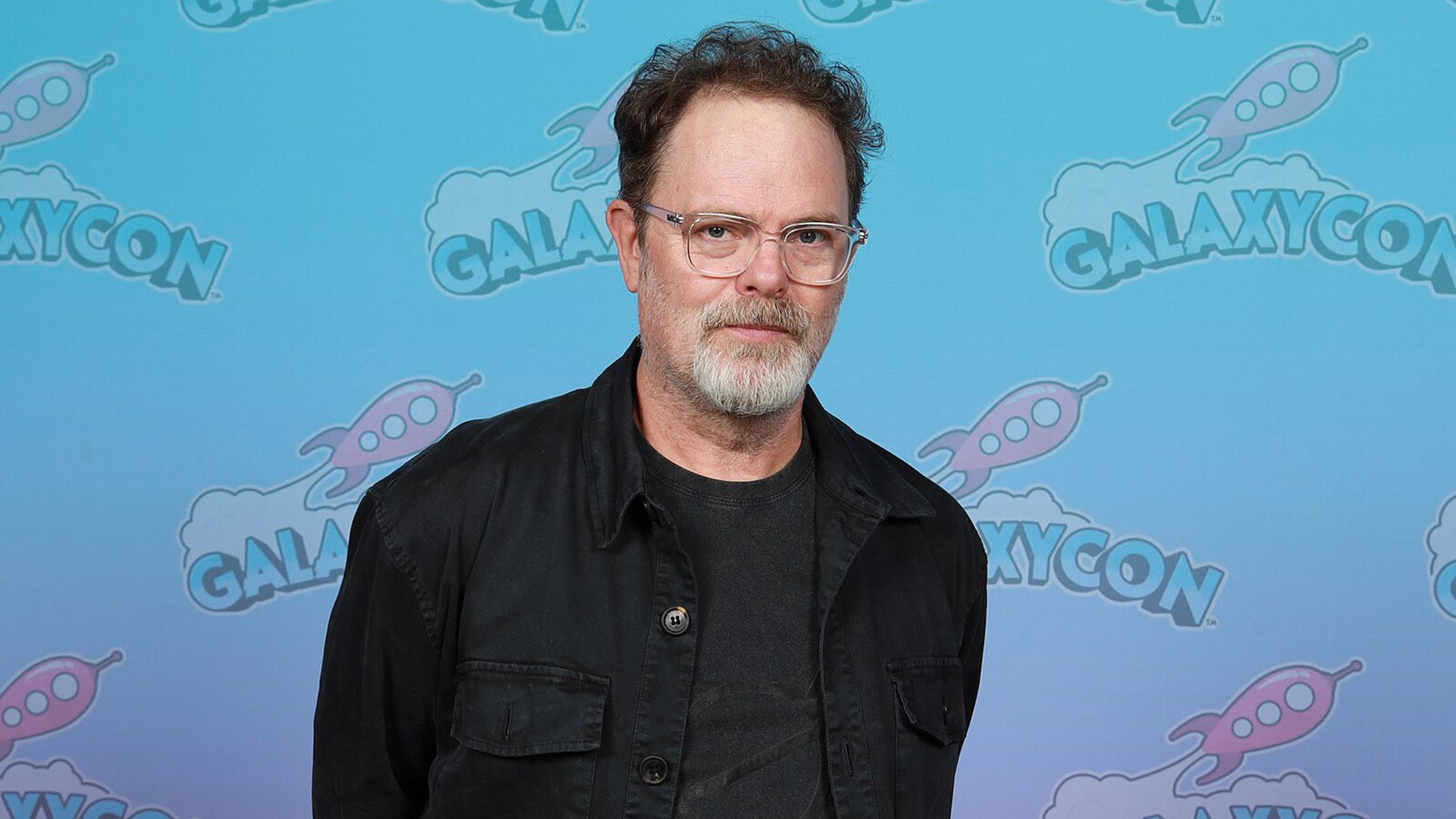 Aktor Rainn Wilson w okularach i czarnej kurtce na niebieskim tle