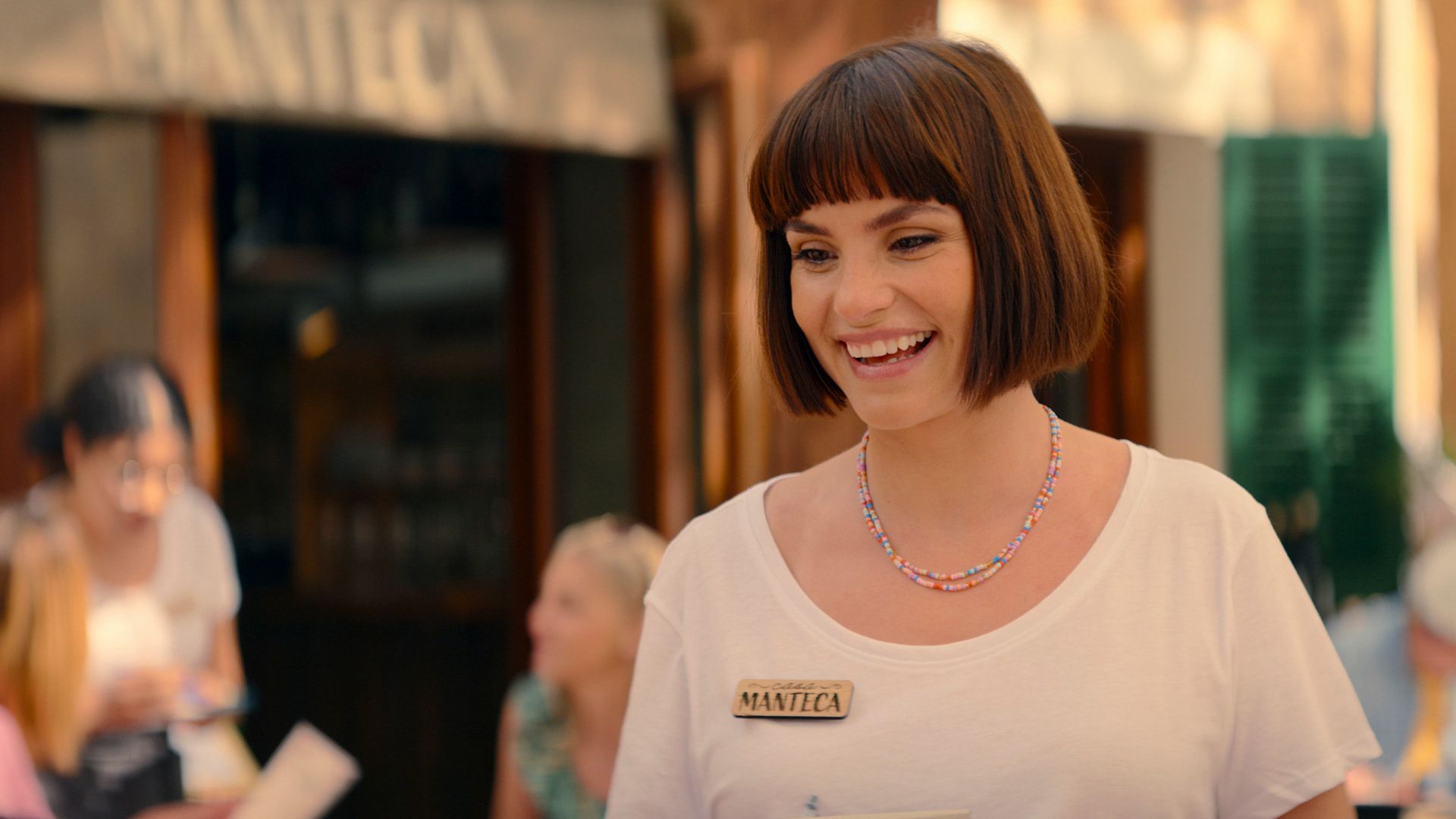 Charlotte Riley z fryzurą bob, ubrana w białą koszulkę, z drewnianą plakietką na piersi i kolorowym naszyjnikiem z koralików