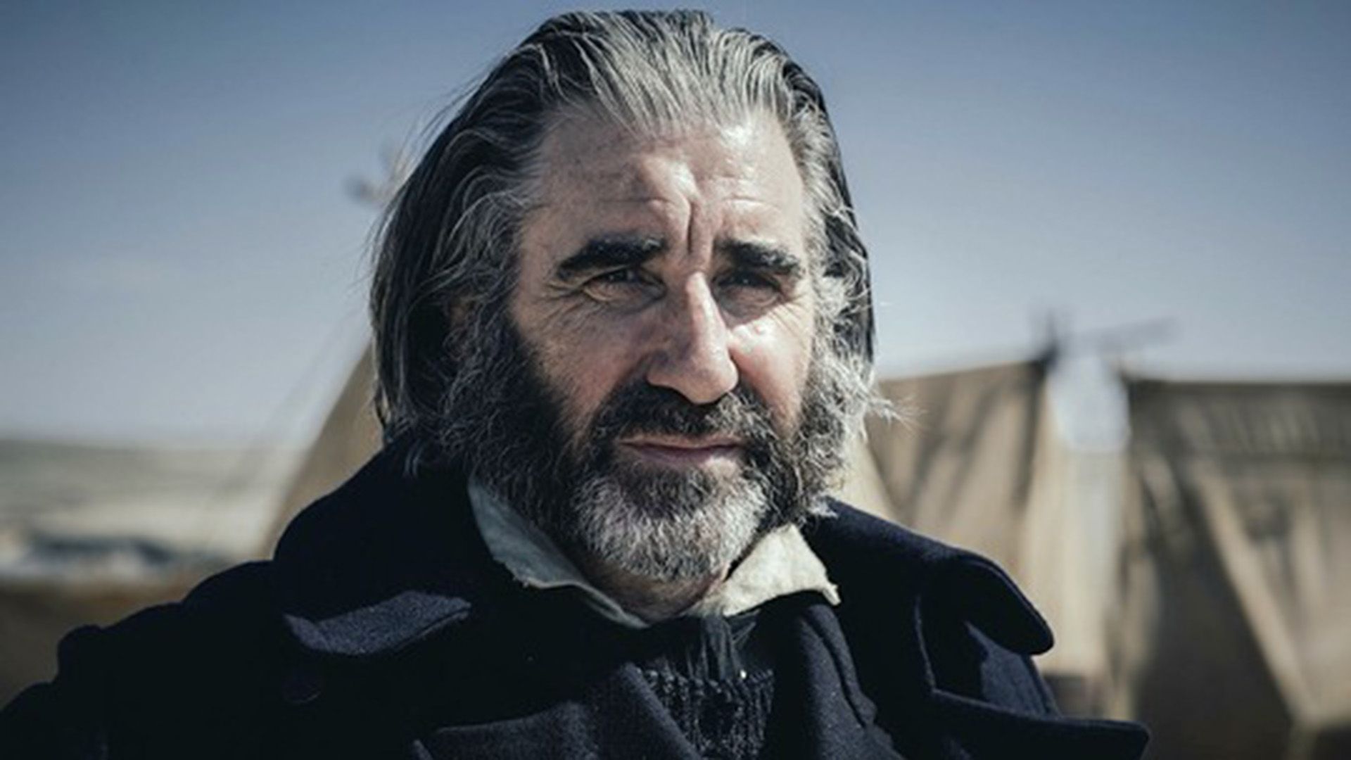 John Lynch z siwą brodą mrużący oczy w słońcu