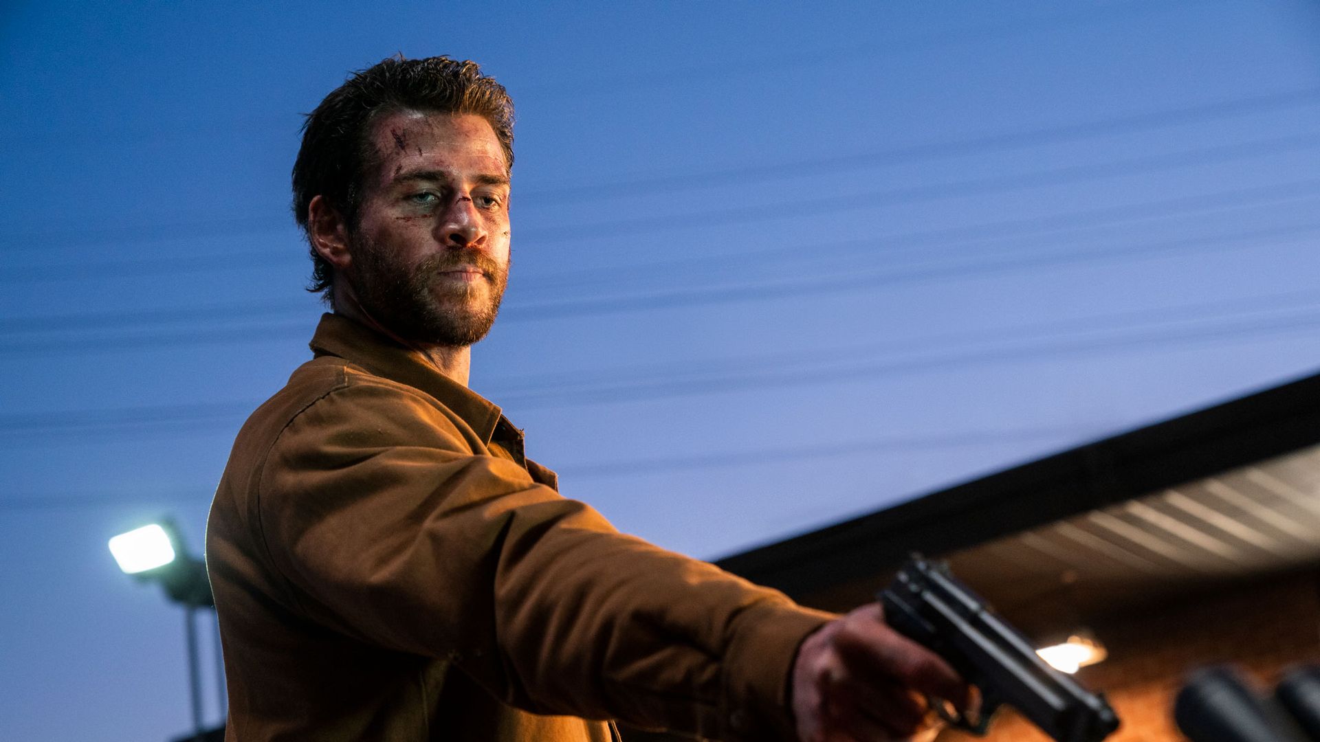 Liam Hemsworth w brązowej kurtce trzymający broń