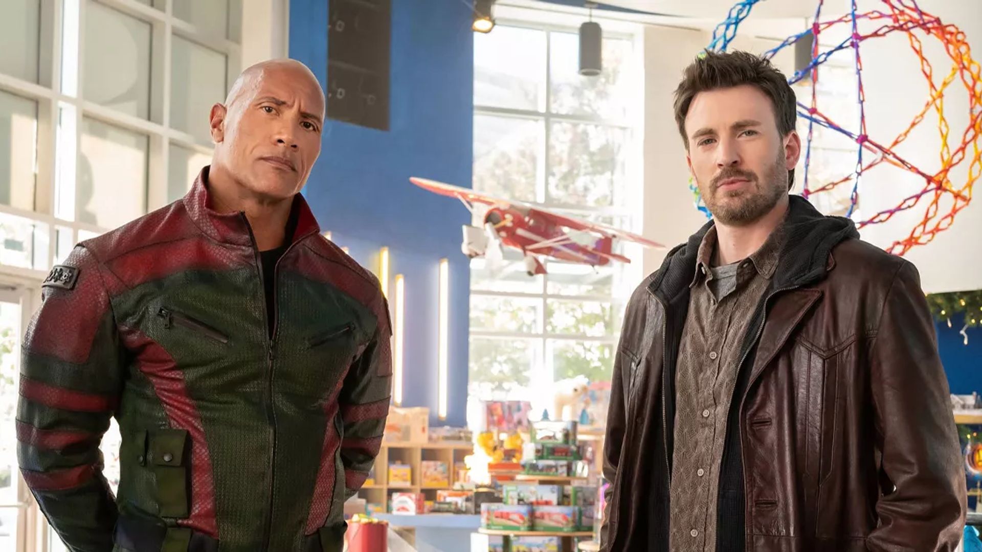 Chris Evans i Dwayne Johnson w kurtkach stoją w jasno oświetlonym pomieszczeniu z zabawkami i modelem samolotu w tle