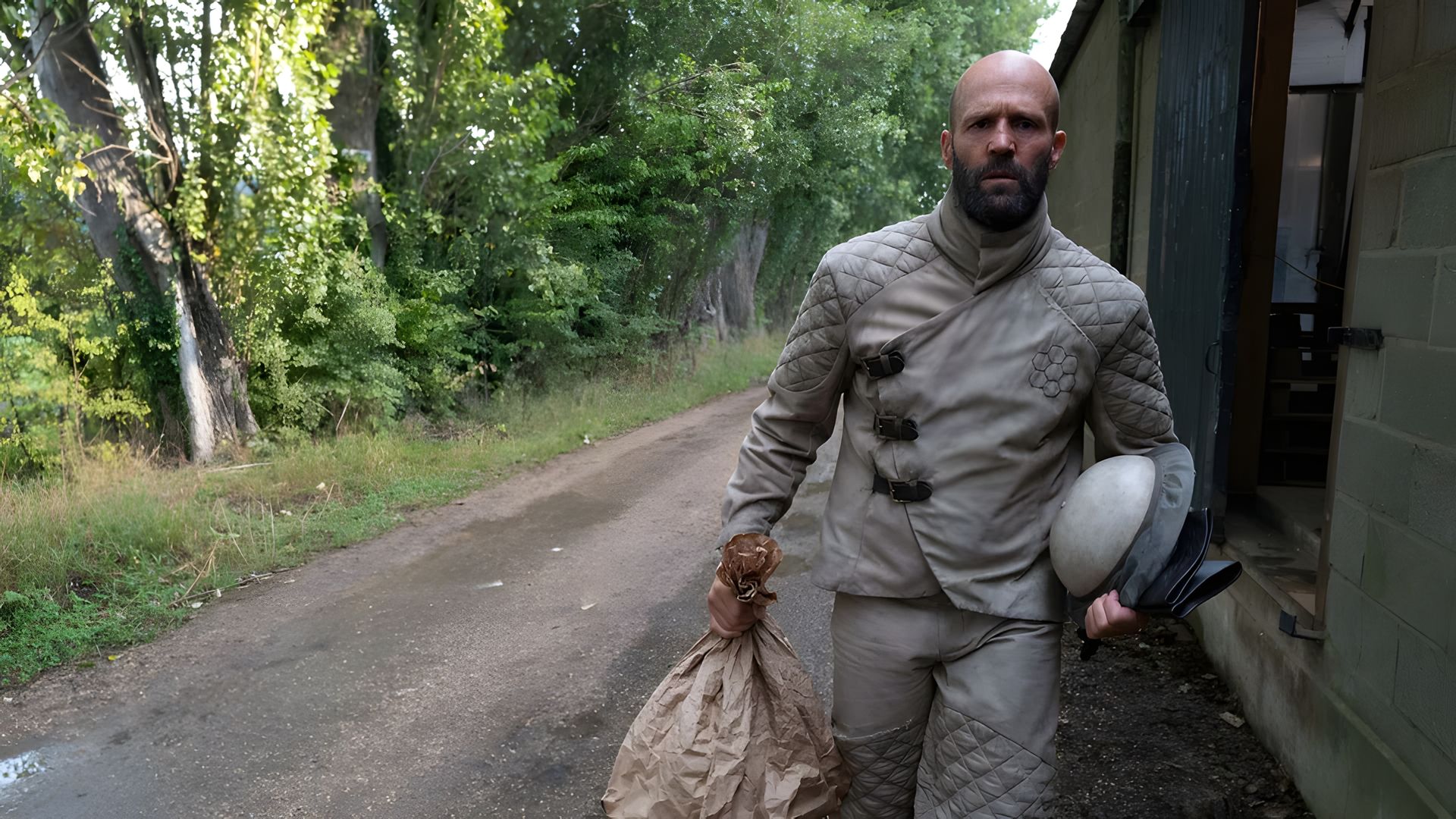Jason Statham w szarym kombinezonie ochronnym idzie drogą niosąc torbę i kask, w tle drzewa