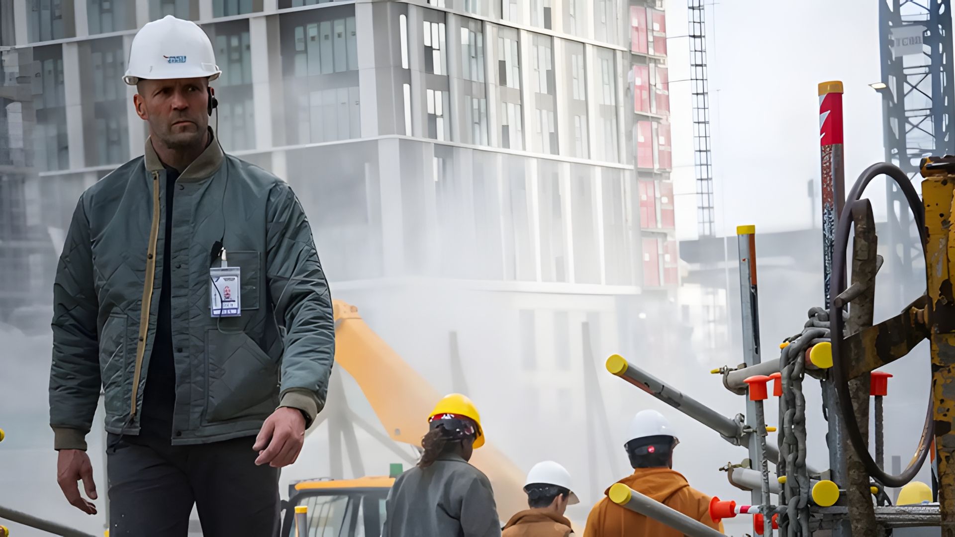 Jason Statham w kasku i kurtce idzie przez plac budowy z widocznymi w zamglonym tle pracownikami i maszynami