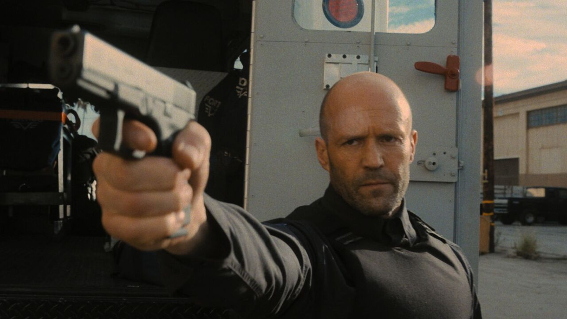 Jason Statham w czarnym stroju taktycznym celuje z pistoletu, stojąc przy furgonetce pancernej na tle przemysłowego krajobrazu