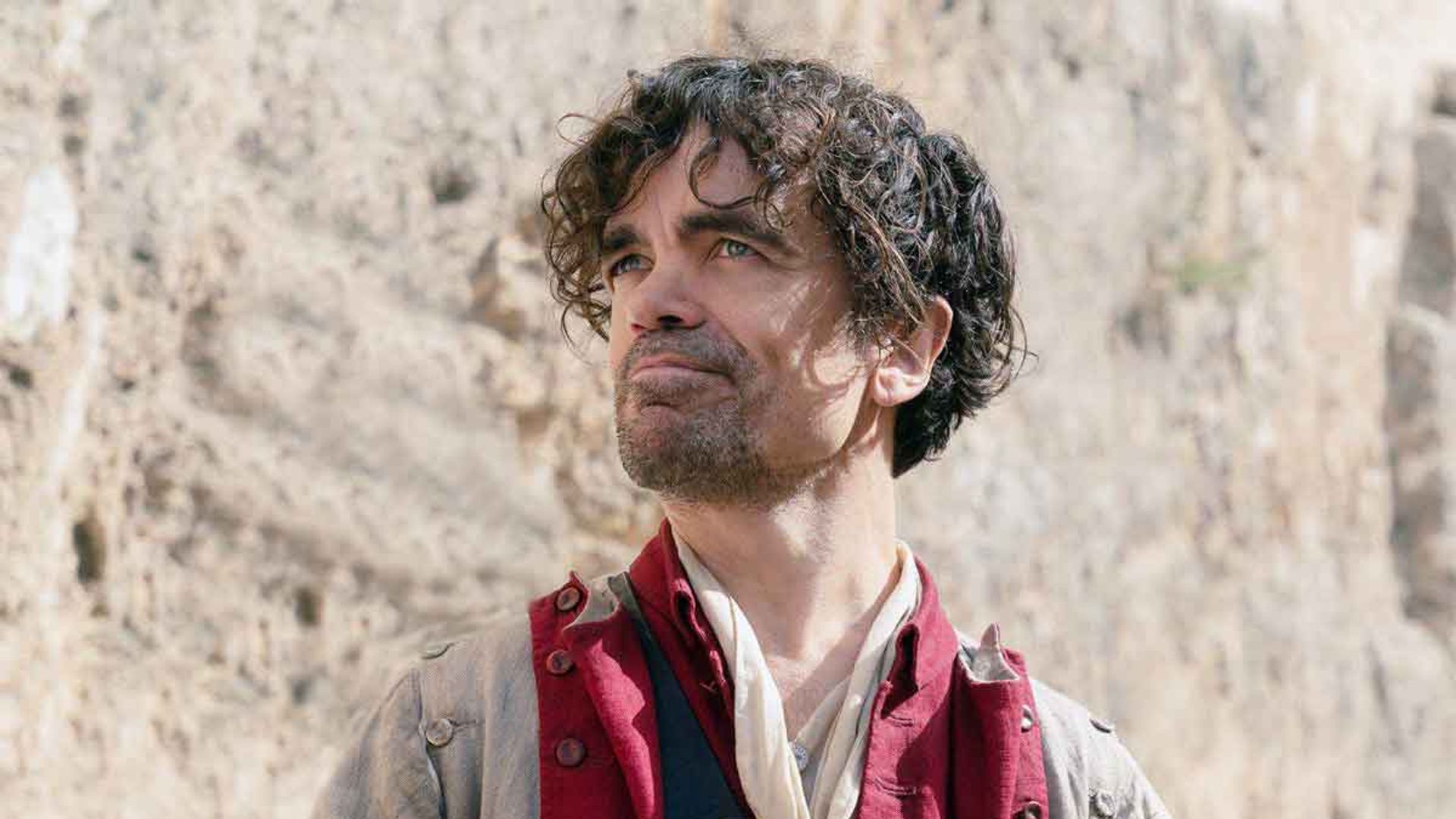 Peter Dinklage w historycznym kostiumie z czerwonym kamizelolem trzyma list i patrzy w górę z lekkim uśmiechem