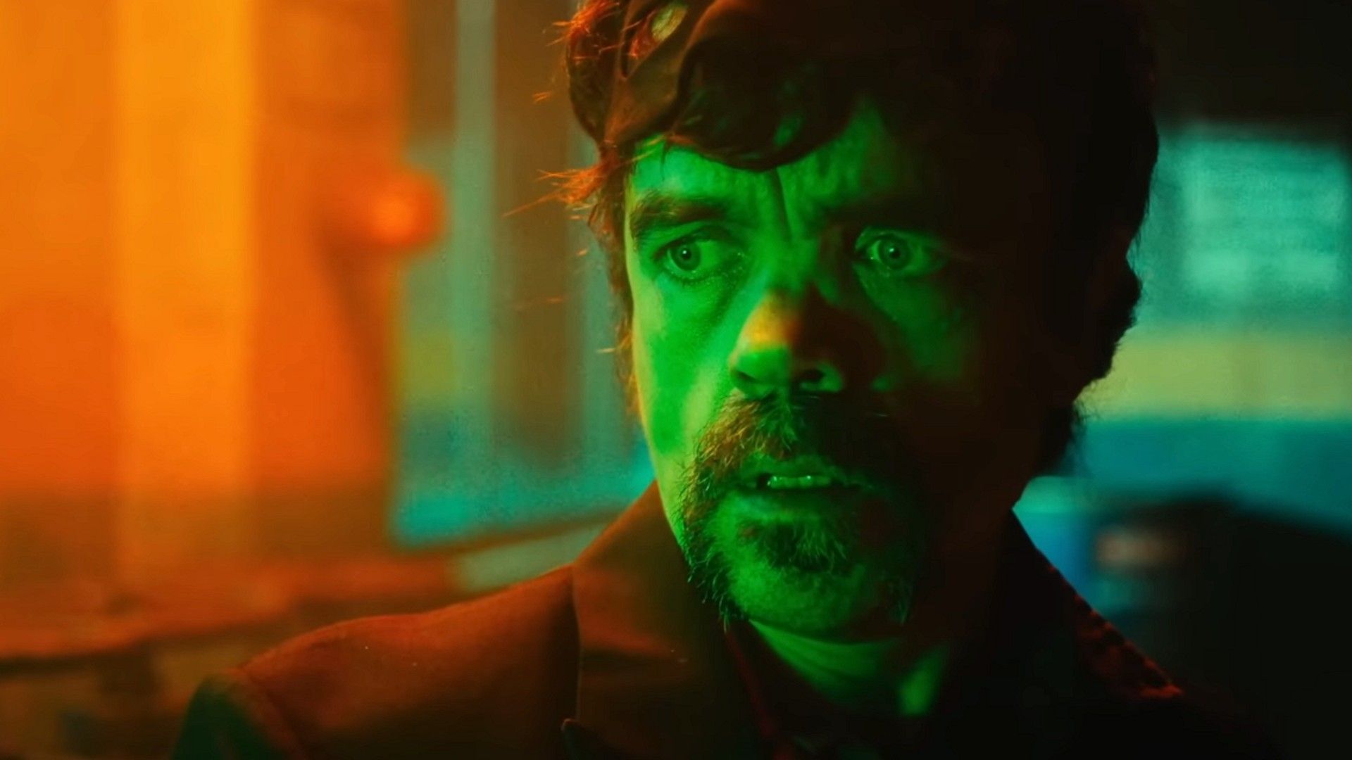 Peter Dinklage stoi w ciemnym pomieszczeniu oświetlonym zielonym i pomarańczowym światłem