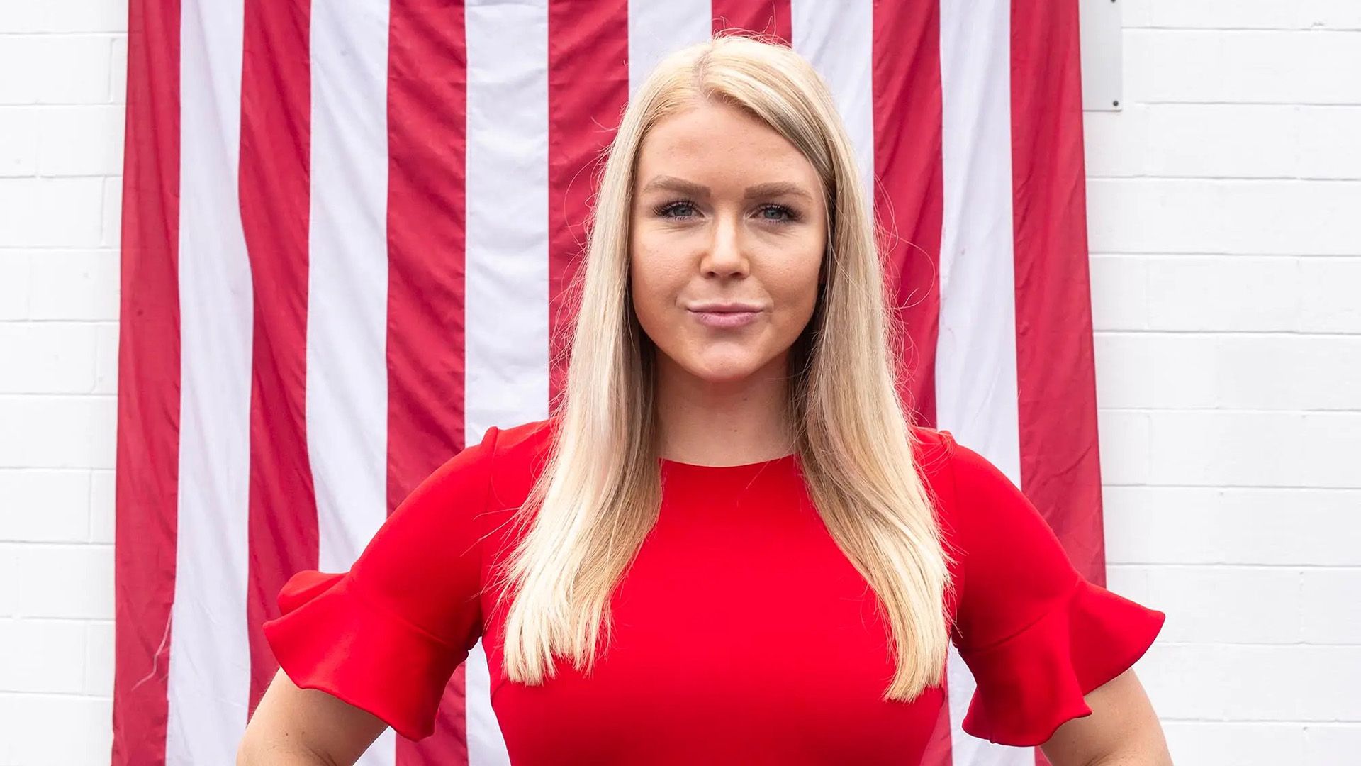 Długowłosa blondynka w czerwonej sukience stoi na tle amerykańskiej flagi