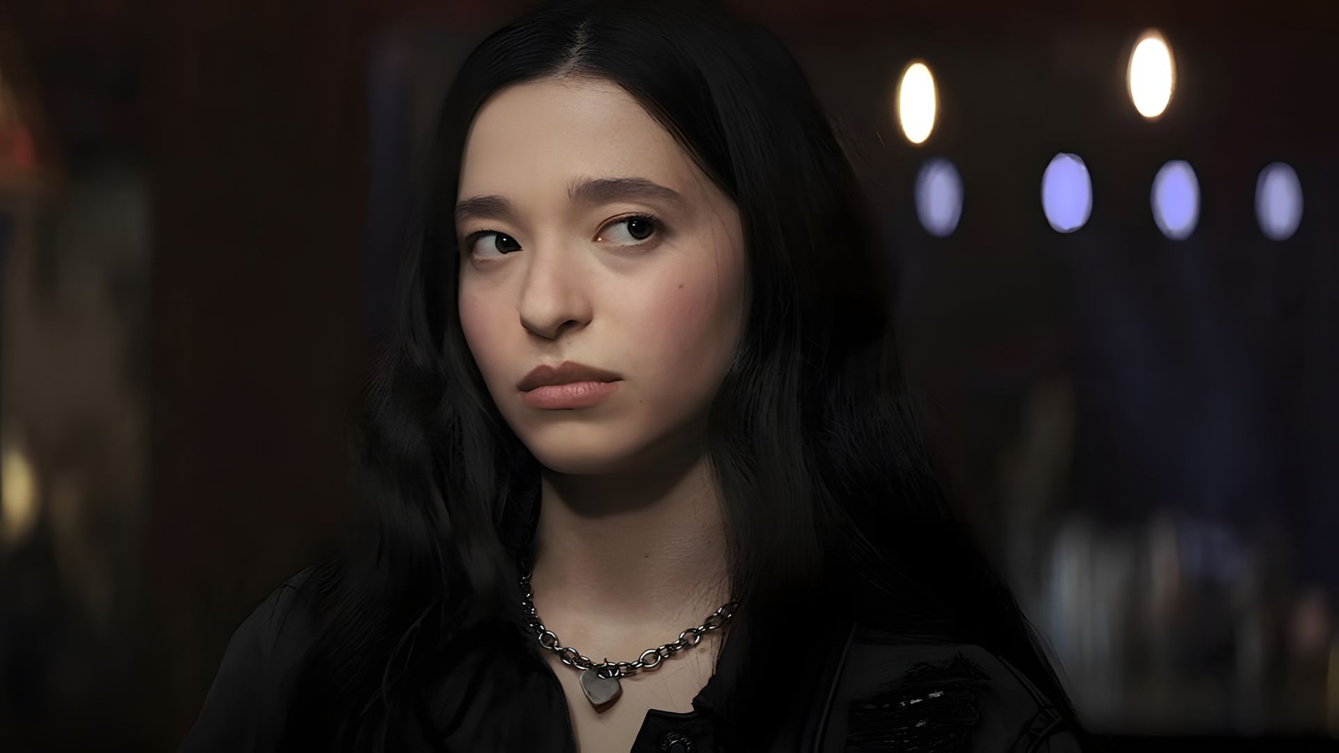 A young girl with long black hair and a chain around her neck is looking to the side with a serious expression on her face, the background is blurred