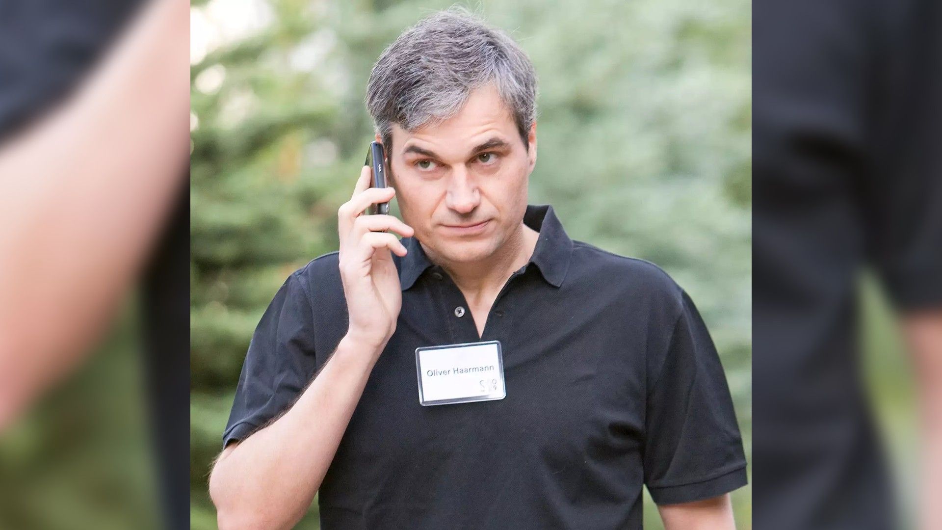 Mężczyzna w czarnej koszuli z plakietką rozmawia przez telefon na tle zielonych drzew, wyglądając na skupionego