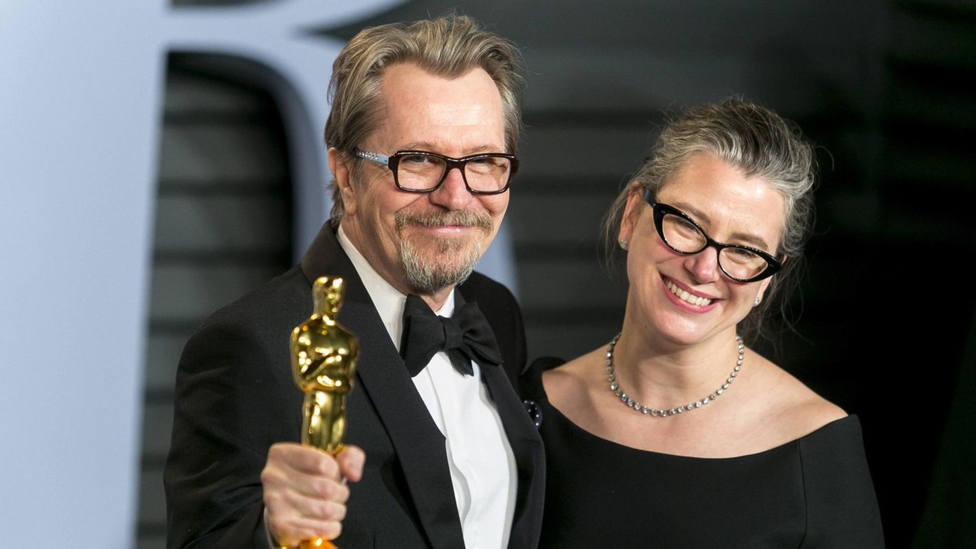 Gary Oldman w smokingu trzyma statuetkę Oscara i uśmiecha się obok Gisele Schmidt w czarnej sukience i okularach