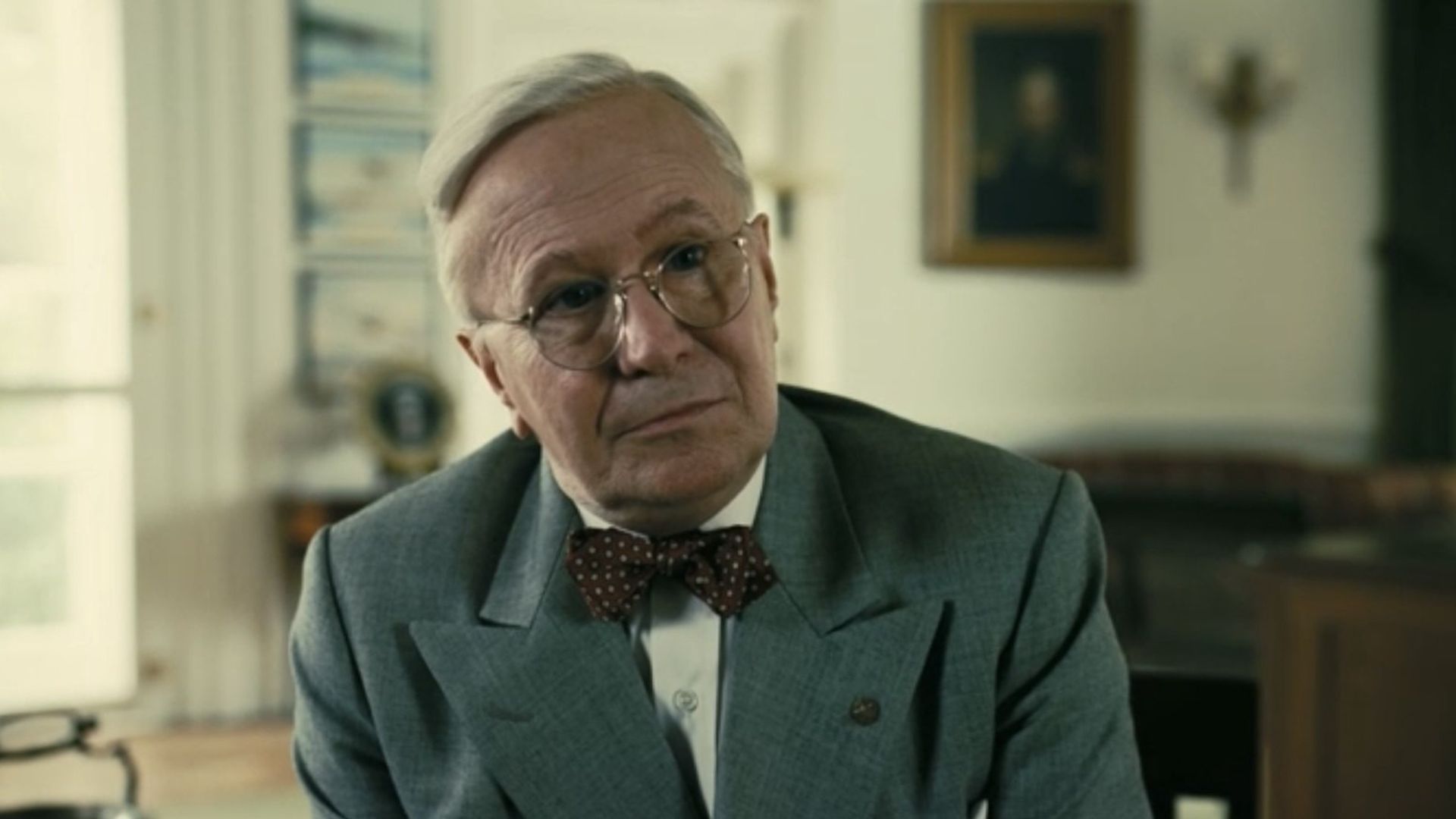 Gary Oldman w okularach i szarym garniturze z muszką siedzi w biurze, patrząc w kamerę z lekkim uśmiechem
