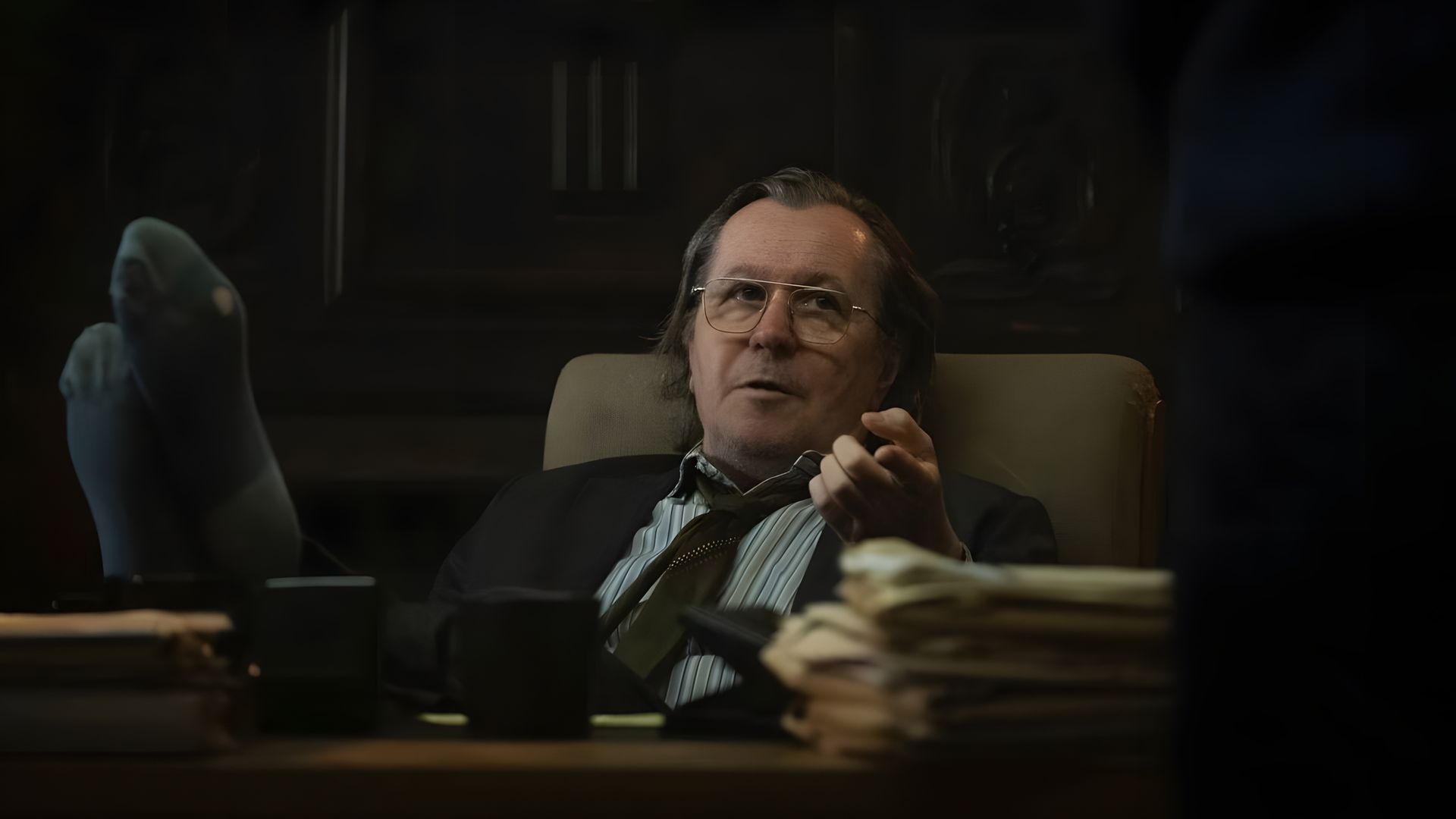 Gary Oldman w okularach i garniturze siedzi przy biurku w biurze z nogami na stole, z zamyślonym wyrazem twarzy
