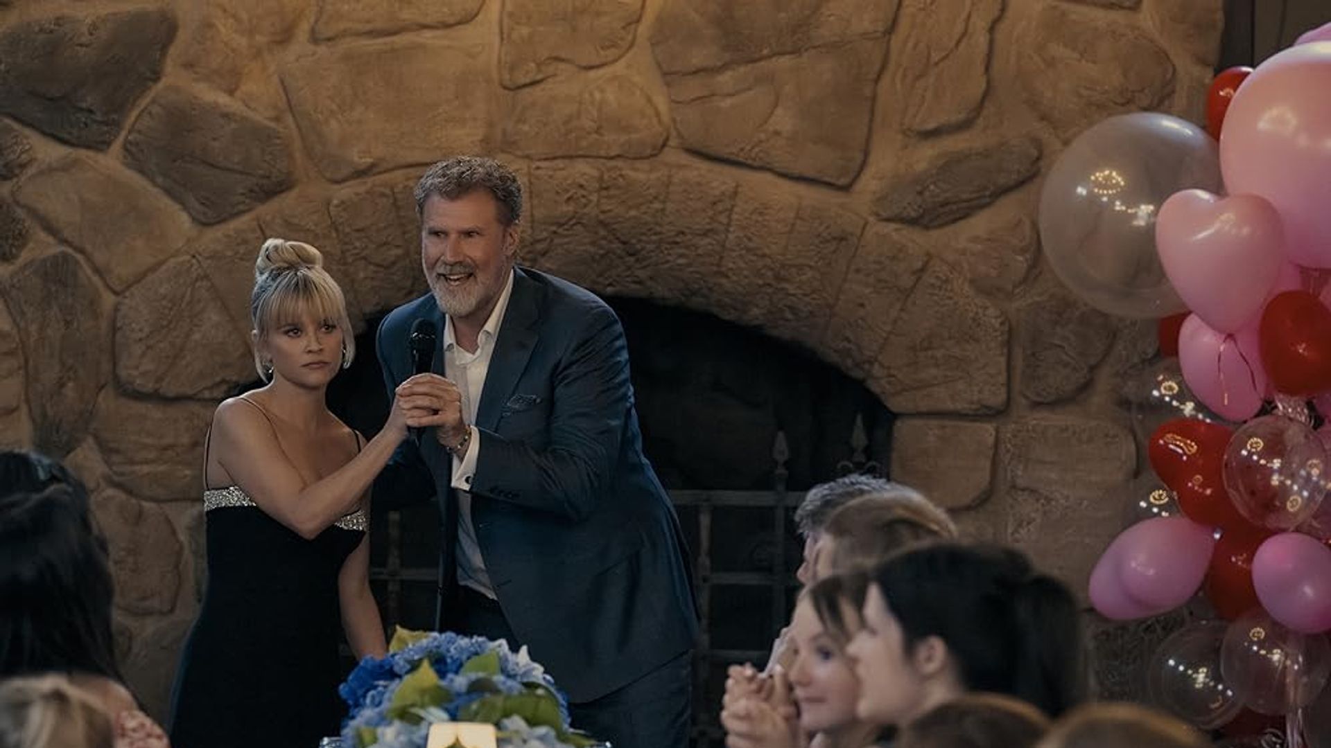 Will Ferrell w garniturze z mikrofonem trzyma za rękę Reese Witherspoon w czarnej sukience na tle kominka, świątecznego stołu i balonów