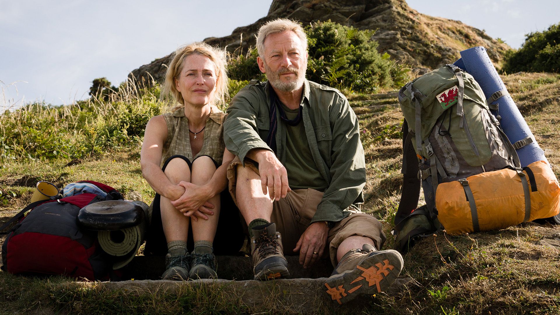Gillian Anderson i Jason Isaacs siedzą na wzgórzu, obok nich leżą plecaki turystyczne