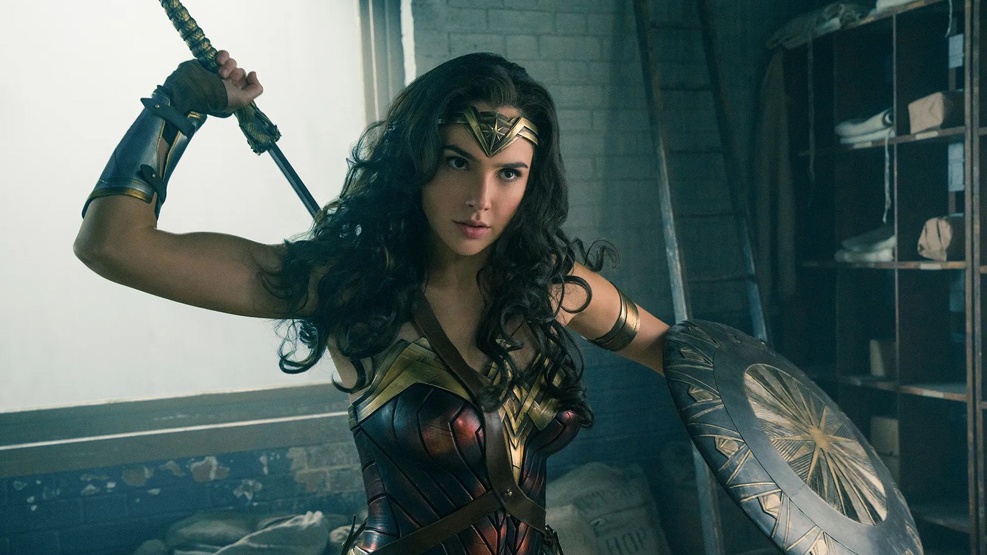 Gal Gadot jako Wonder Woman: w zbroi, trzymająca tarczę lewą ręką, wyciągająca miecz zza pleców prawą ręką