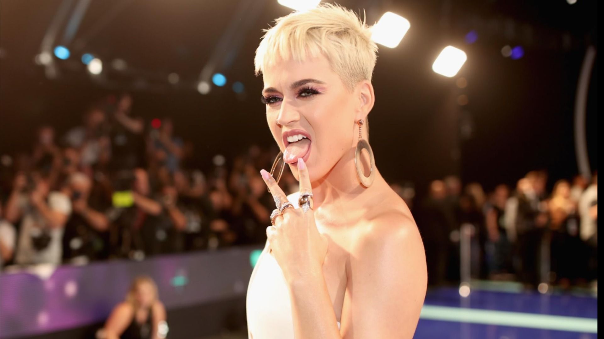 Piosenkarka Katy Perry wystawia język i pokazuje dwa palce w białej sukience na tle publiczności