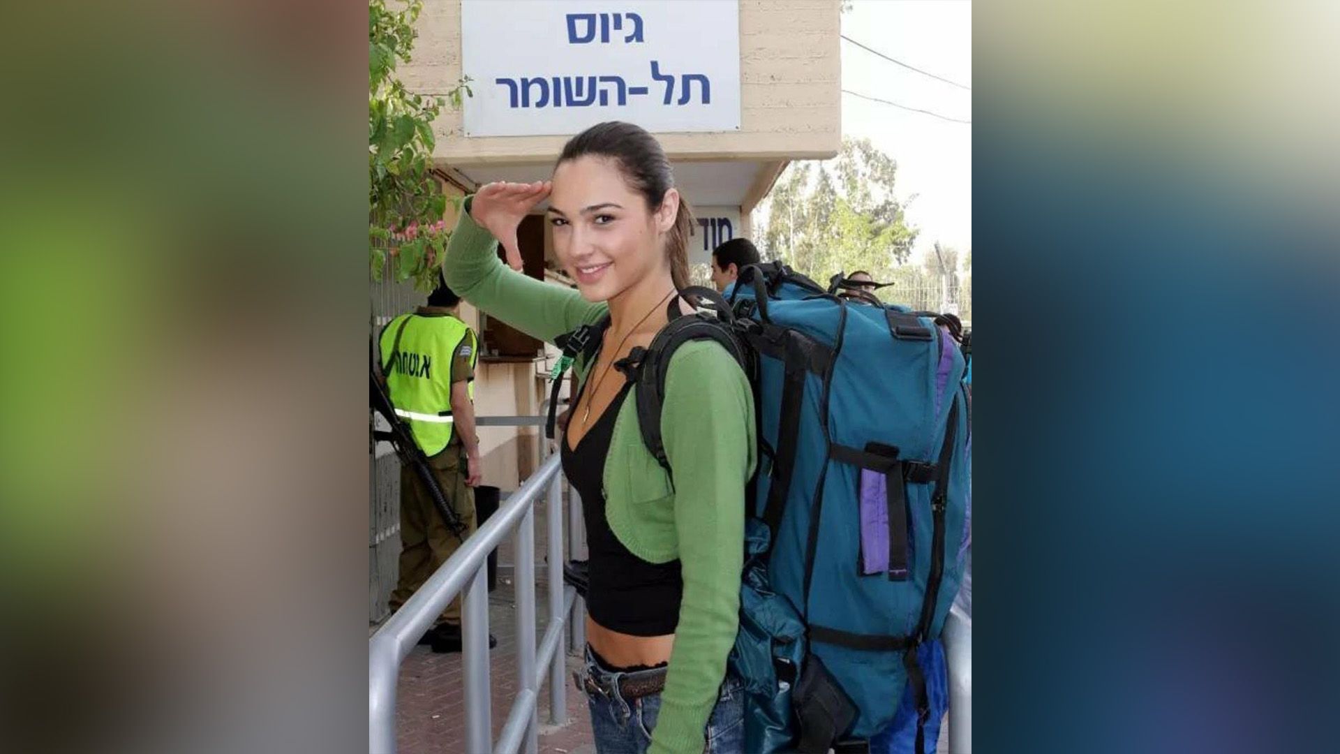 Młoda Gal Gadot w zielonym swetrze i z dużym niebieskim plecakiem na plecach salutuje prawą ręką
