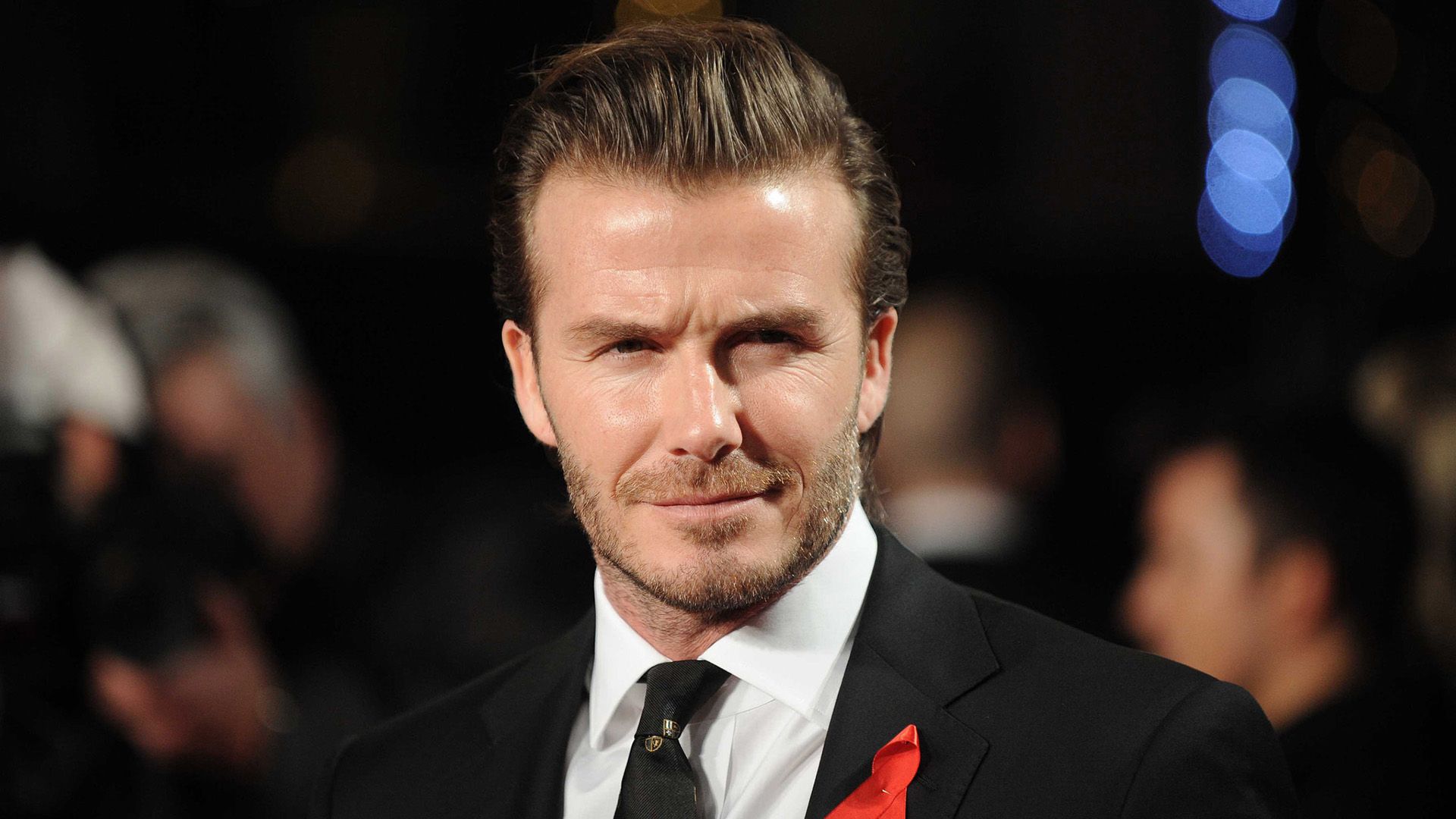Piłkarz David Beckham w czarnej kurtce z przypiętą czerwoną wstążką