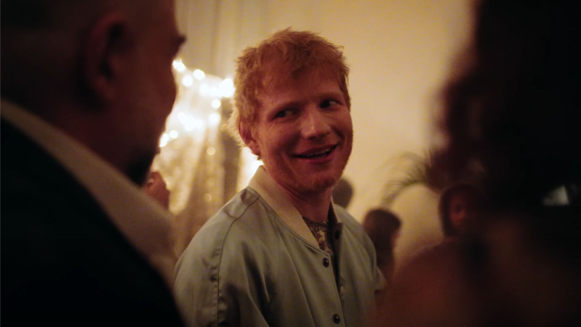 Piosenkarz Ed Sheeran patrzy i uśmiecha się w żółto-pomarańczowym świetle obok sylwetek ludzi