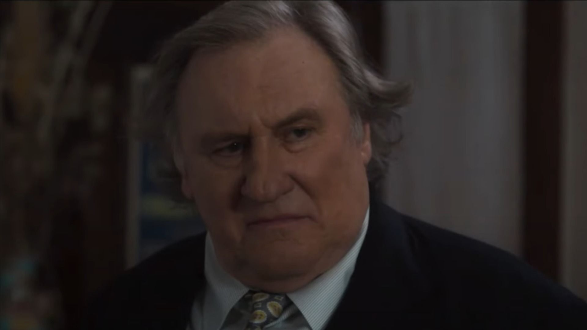 Aktor Gérard Depardieu w klasycznym garniturze patrzy w bok na rozmyte tło