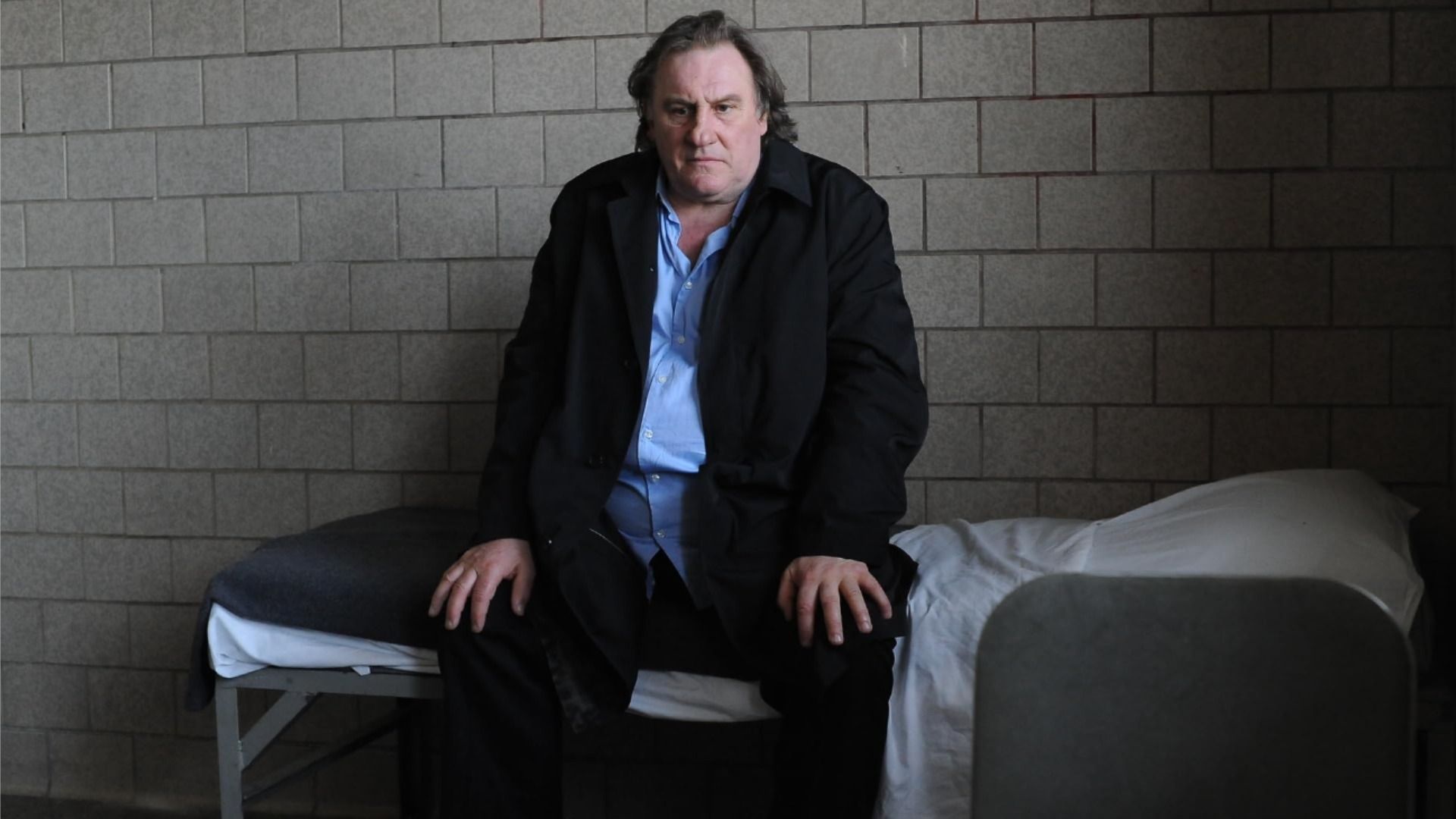 Aktor Gérard Depardieu w klasycznym garniturze siedzi na małym łóżku i patrzy na podłogę