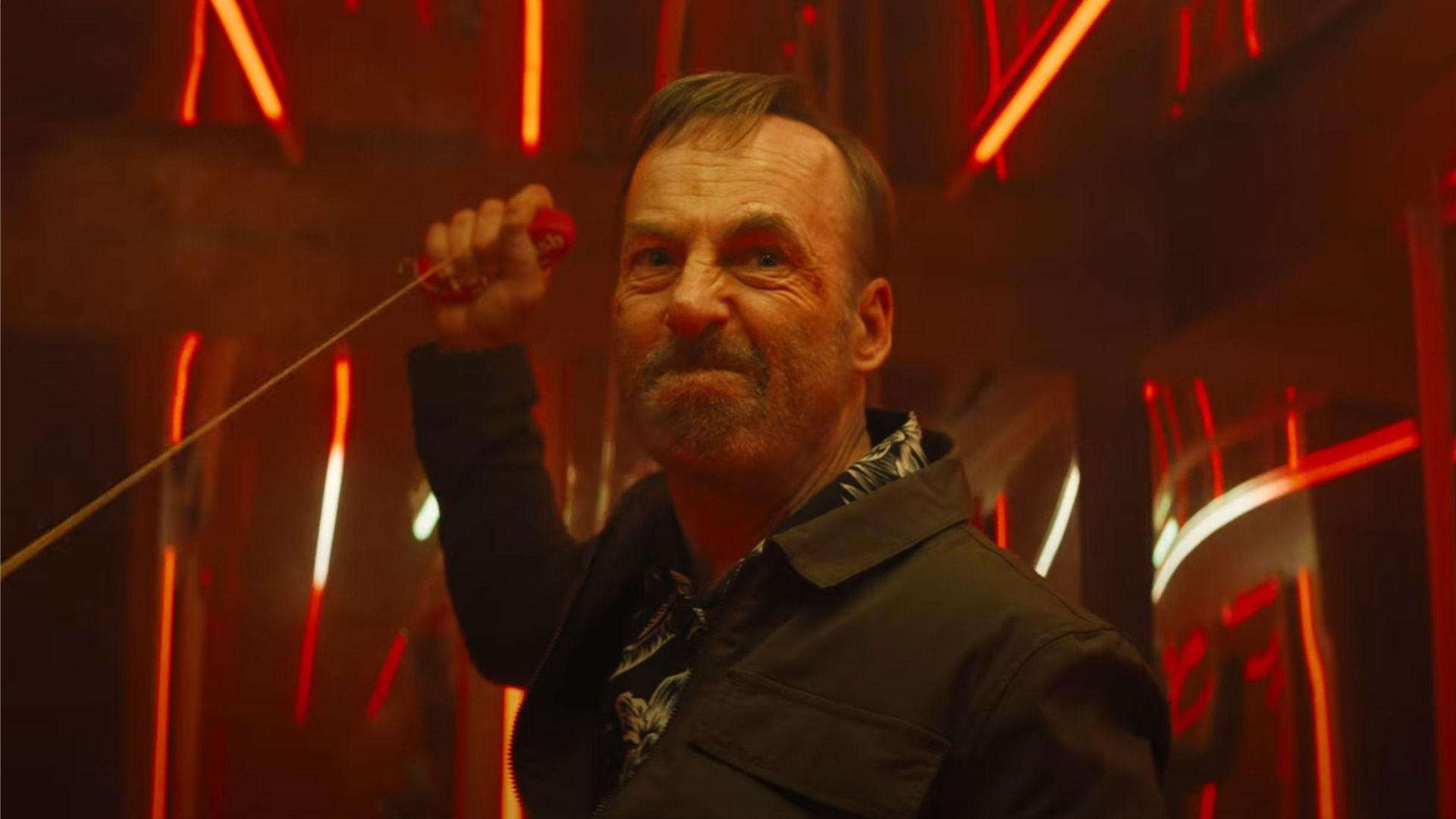 Aktor Bob Odenkirk w czarnej kurtce trzyma linę w rękach i patrzy gniewnie w kierunku czerwonego światła