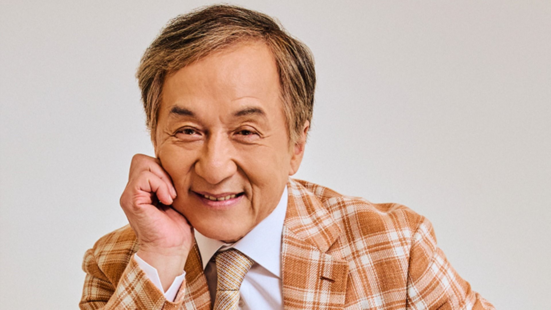 Aktor Jackie Chan w kraciastej brązowej marynarce siedzi, opierając się na ręce, i uśmiecha się do kamery na białym tle