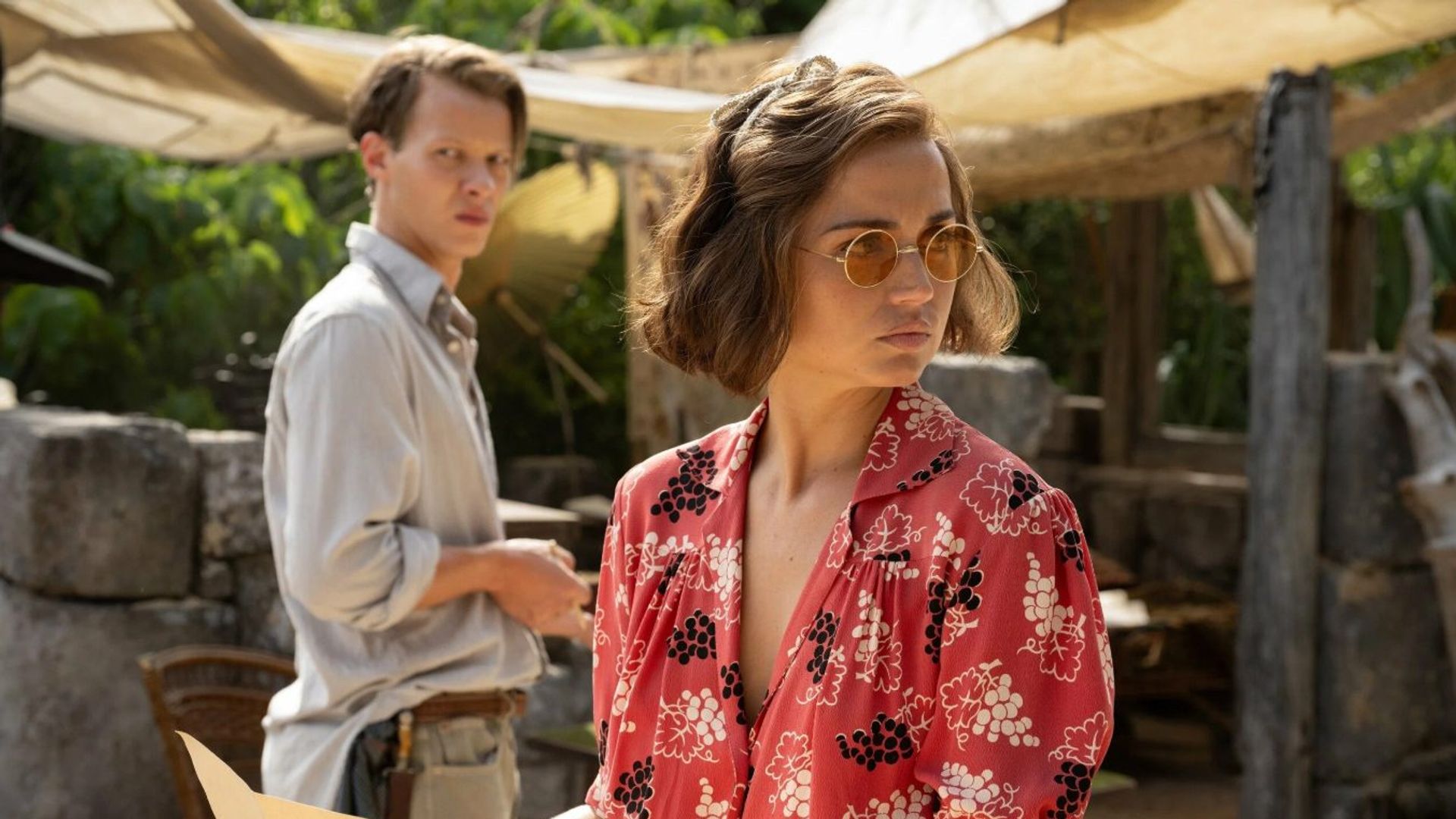 Ana de Armas w czerwonej, wzorzystej bluzce i okularach przeciwsłonecznych trzyma mapę, stojąc obok mężczyzny w jasnej koszuli, scena osadzona na tle ruin archeologicznych