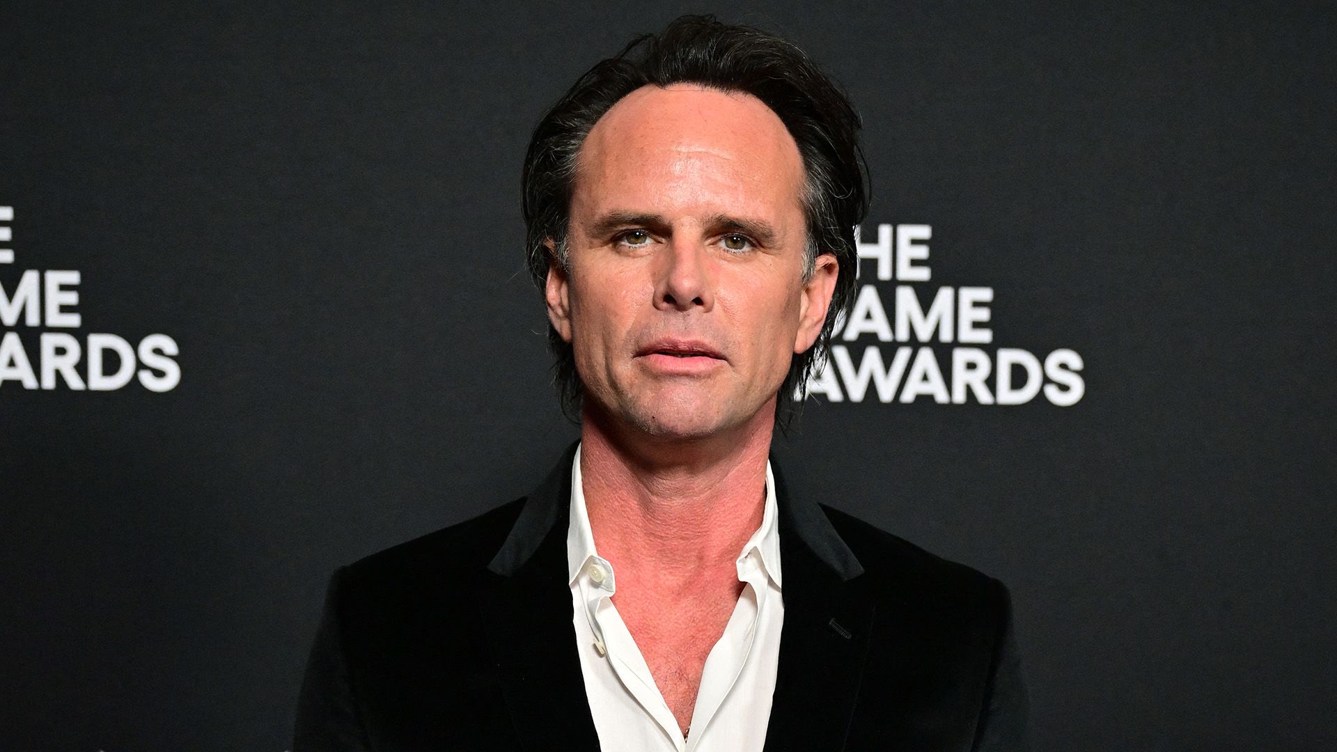 Aktor Walton Goggins w czarnej marynarce i białej koszuli z rozpiętym kołnierzem na czarnym tle