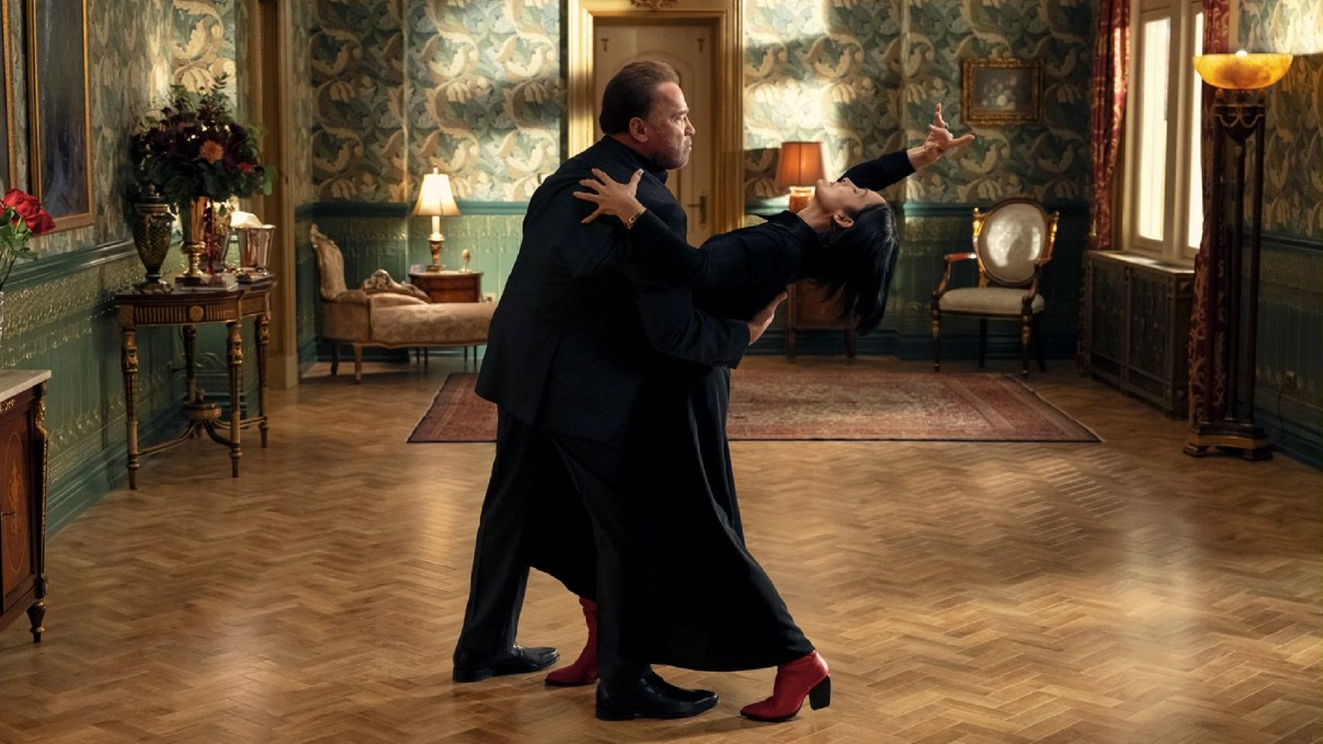 Arnold Schwarzenegger i Carrie-Anne Moss tańczący w czarnych garniturach pośrodku pokoju