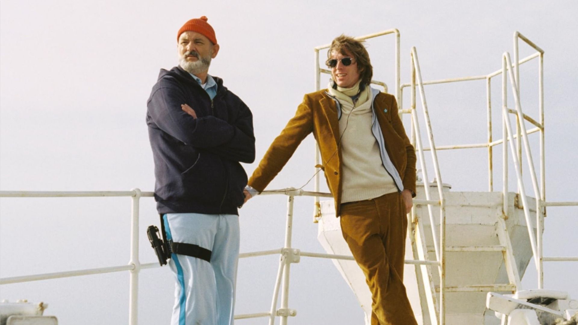 Aktor Bill Murray w czerwonym kapeluszu i reżyser Wes Anderson w brązowej kurtce trzymają się poręczy statku na tle nieba
