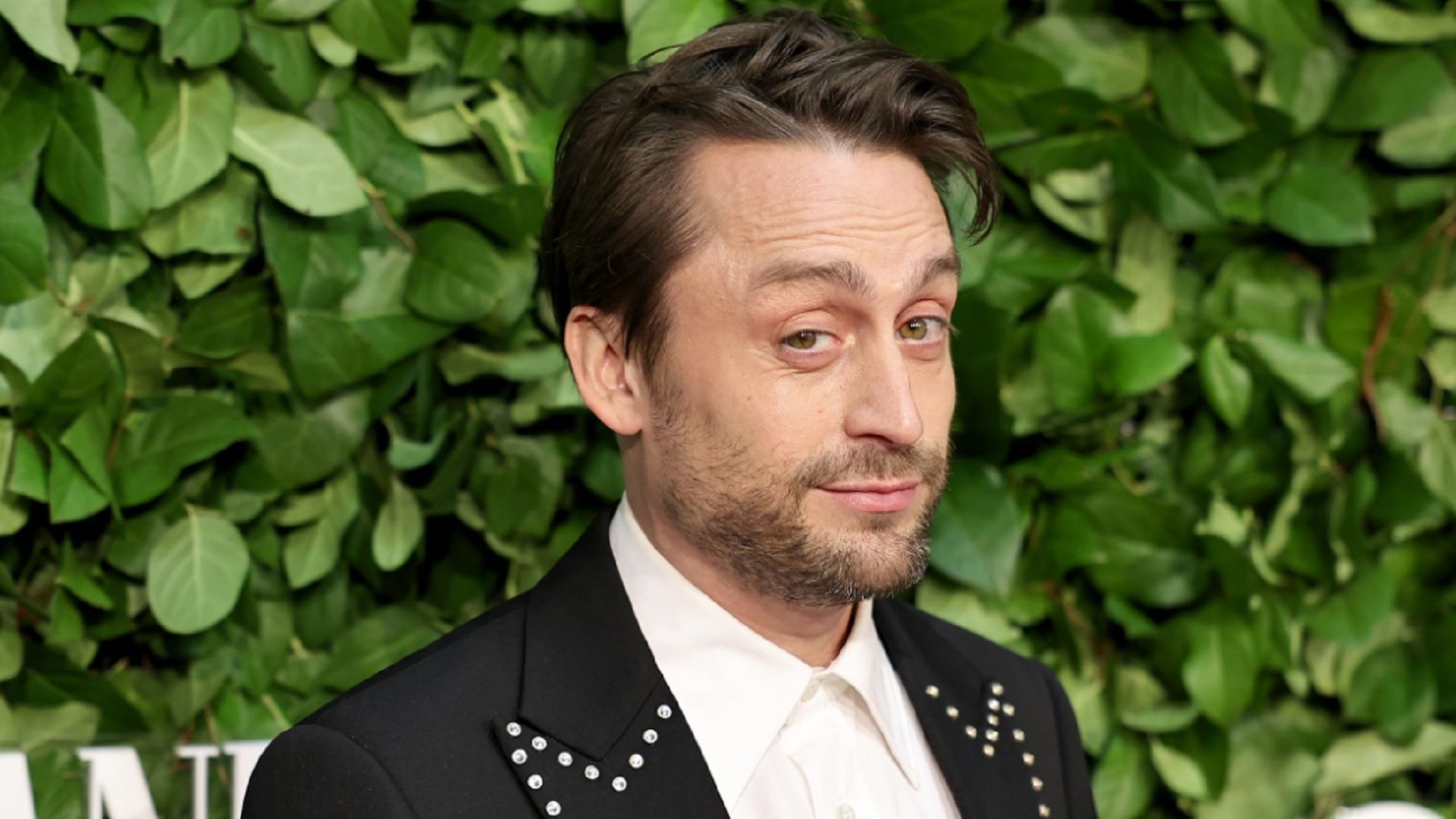 Kieran Culkin patrzący ironicznie w kamerę na tle ściany z liści