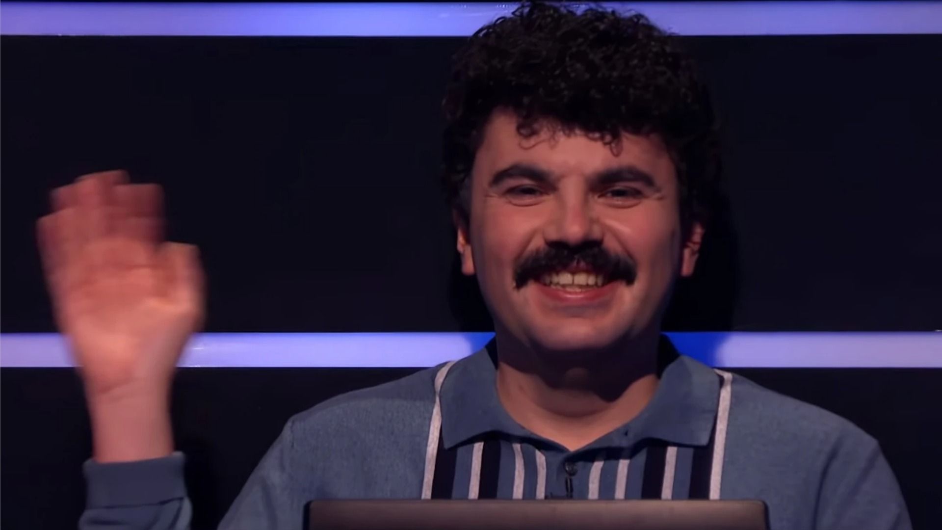 A man in a gray sweater with black stripes and a mustache smiles at the camera and waves his hand against a dark background