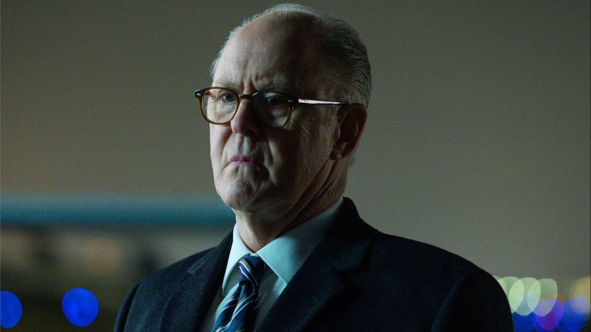 Aktor John Lithgow w garniturze i okularach stoi bez emocji i patrzy w bok