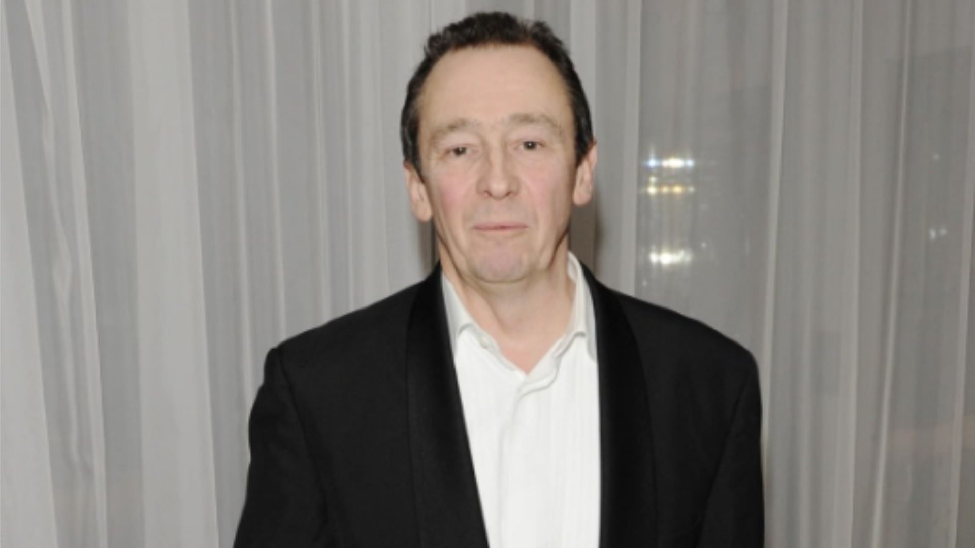 Aktor Paul Whitehouse stoi w czarnym garniturze na tle białej tiulu i patrzy w kamerę