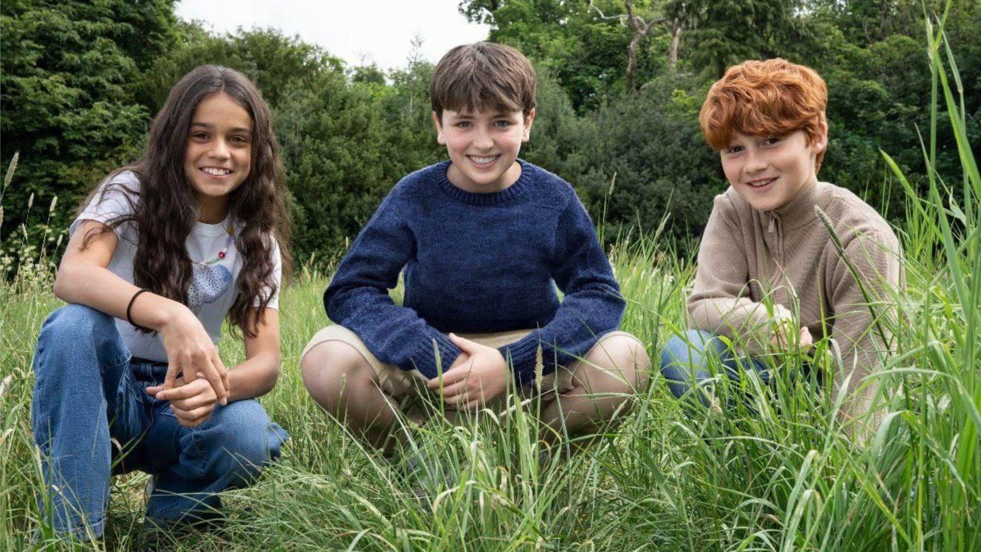Aktorzy Dominic McLaughlin, Alastair Stout i Arabella Stanton siedzą na trawie i uśmiechają się