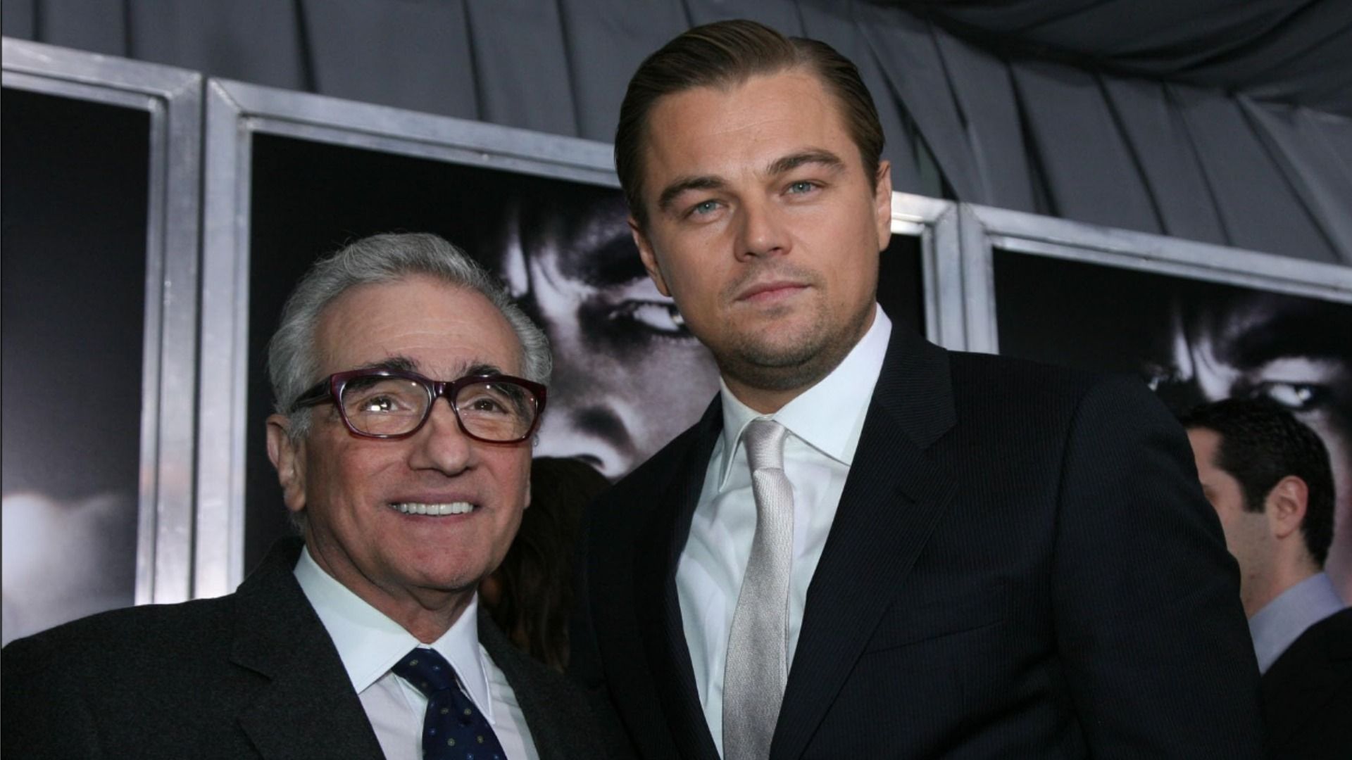 Reżyser Martin Scorsese w okularach stoi i uśmiecha się obok aktora Leonardo DiCaprio, obaj w czarnych garniturach na tle plakatów