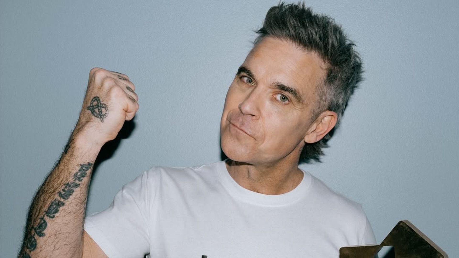 Piosenkarz Robbie Williams uniósł pięść i uśmiecha się, patrząc w kamerę na szarym tle