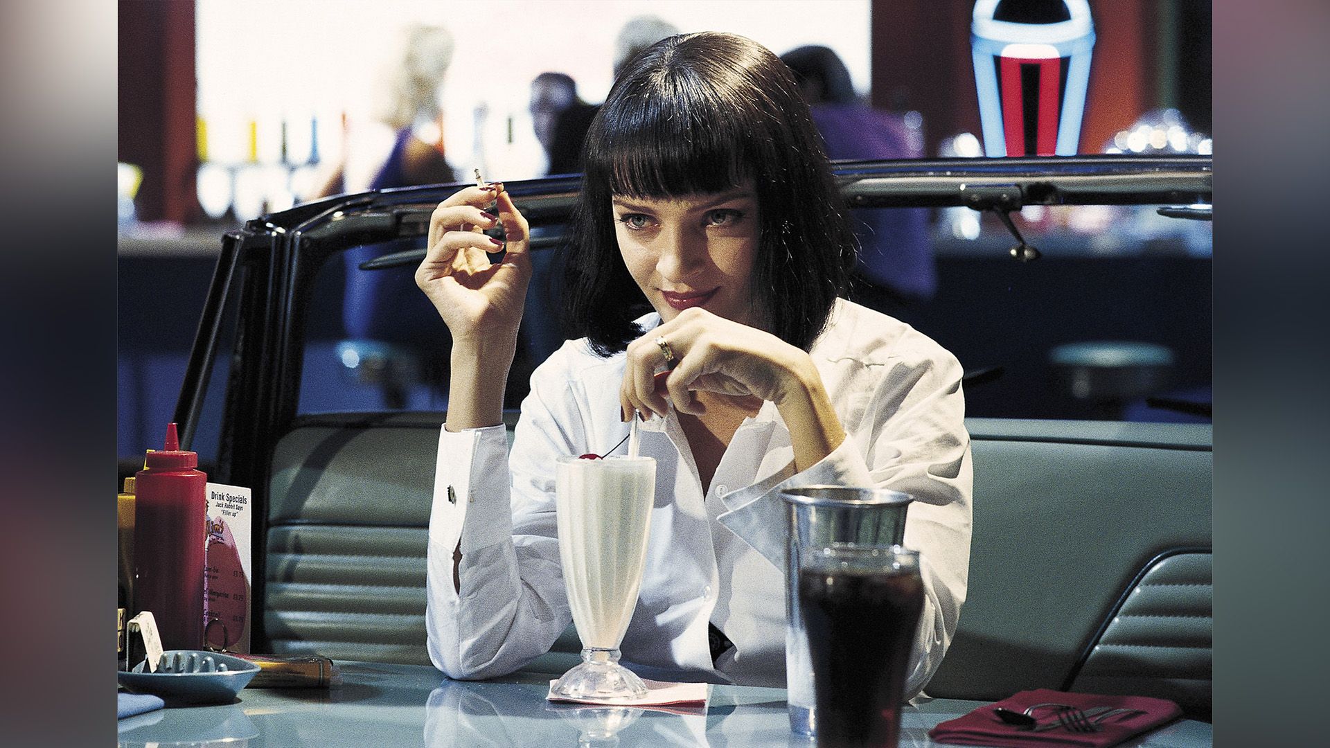 Uma Thurman w białej koszuli pije mleczny koktajl w kawiarni w scenie z filmu 'Pulp Fiction'