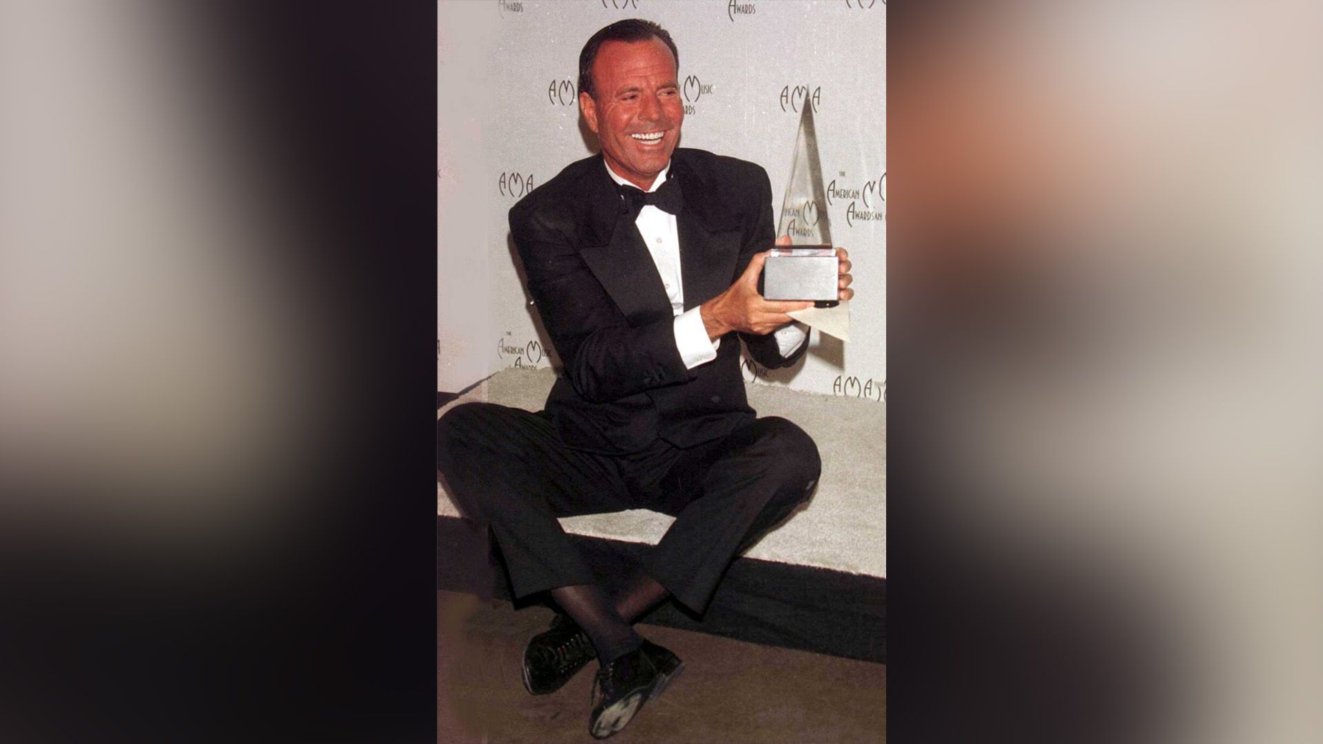 Szczęśliwy Julio Iglesias w czarnym garniturze trzyma w rękach trofeum American Music Awards