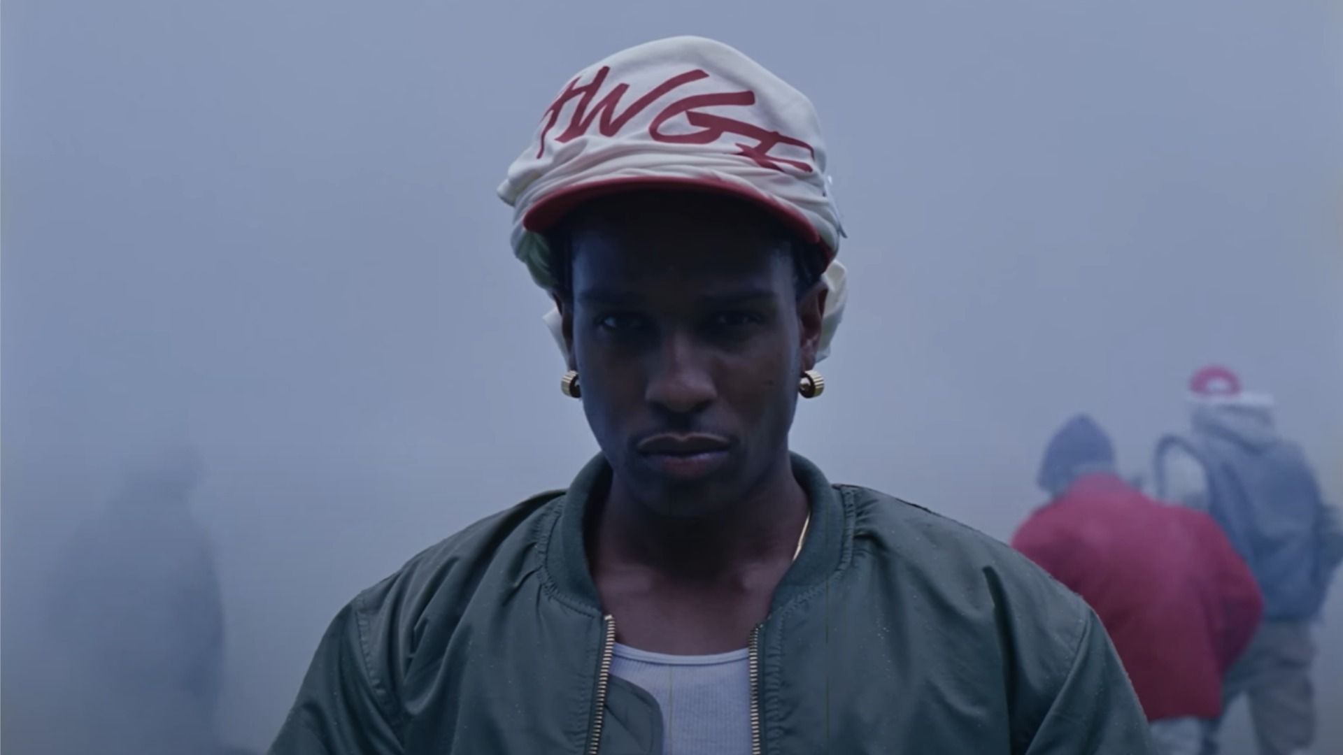Raper A$AP Rocky w zielonej kurtce bomberce patrzy w kamerę na tle zadymionej ulicy